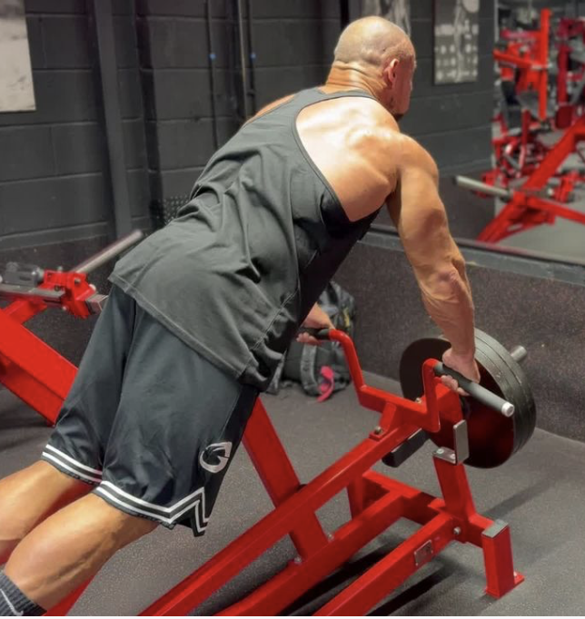A man with a bald head and muscular arms using a push sled with weights at the gym, pushing forward.