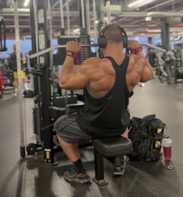 Man with muscular back wearing headphones, black tank top, and shorts, working out on a pull-up machine at the gym.
