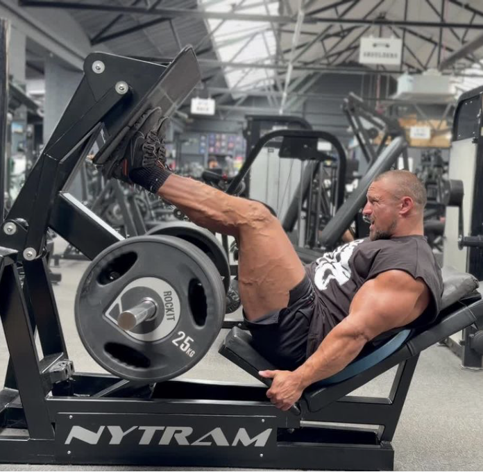 Man performing leg press exercise in a gym, lying on a leg press machine with weight plates, in a spacious gym with various equipment and a high ceiling.