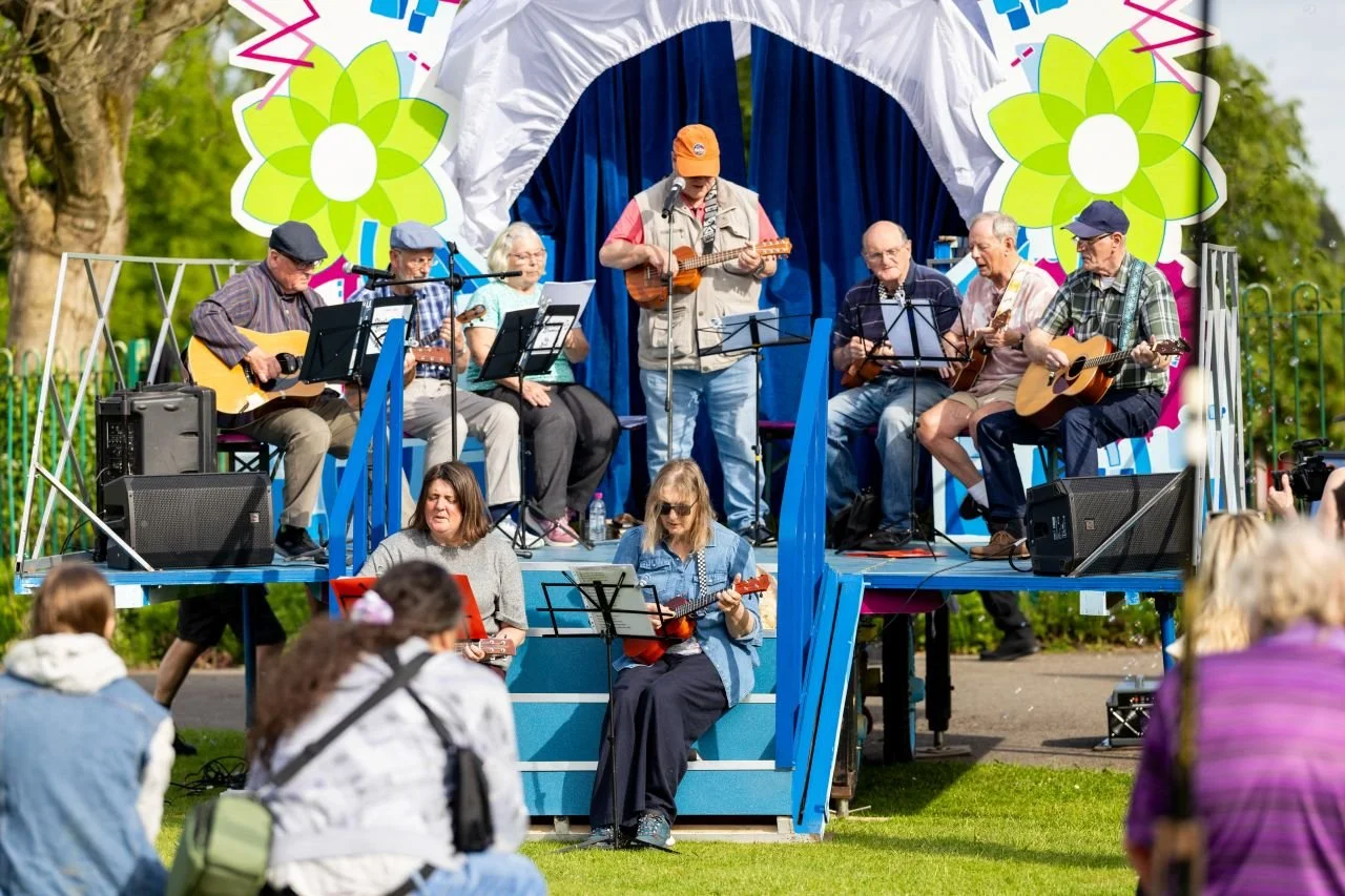 A group of people playing guitars and singing on an outdoor stage during a festival, with an audience seated on the grass in front of them.