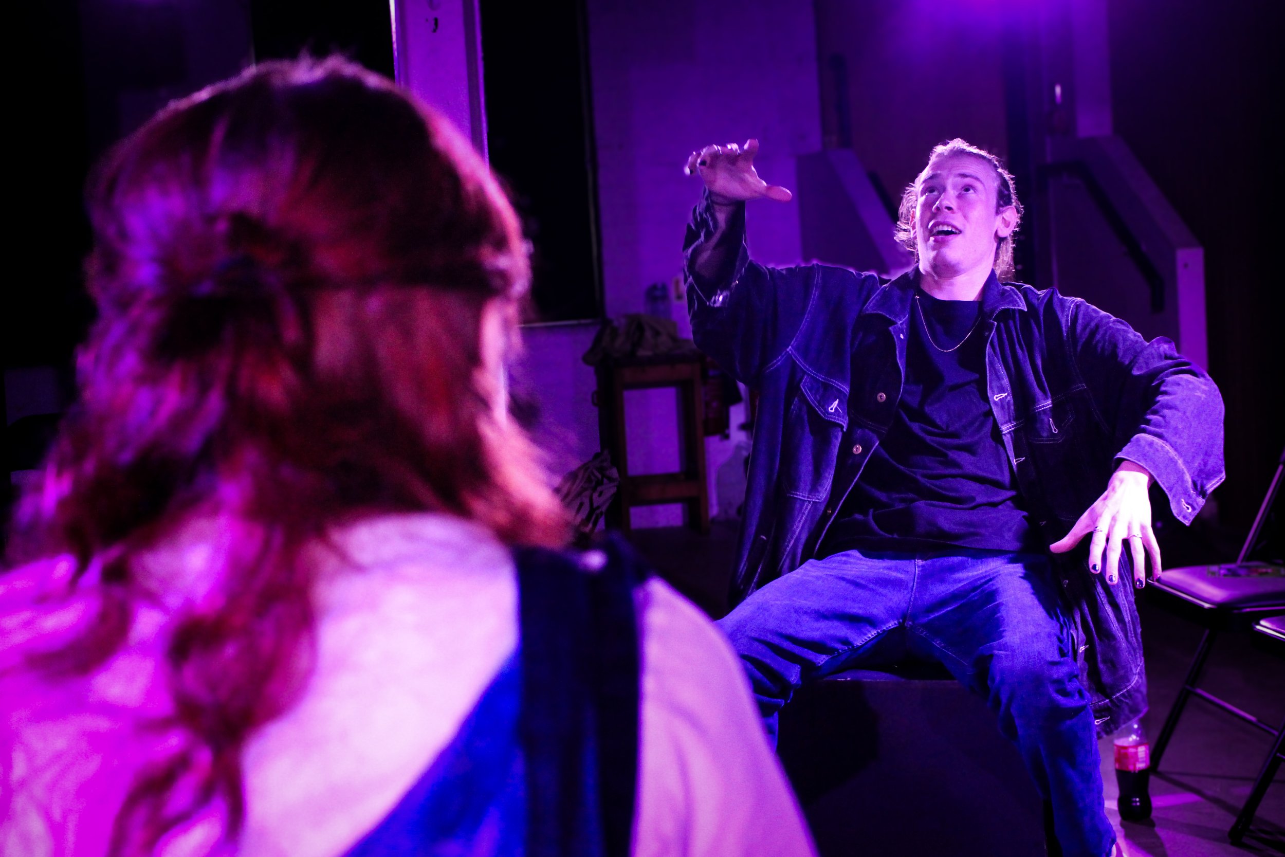 A young man with long hair speaking or singing in a purple-lit room, with a person with red hair in the foreground