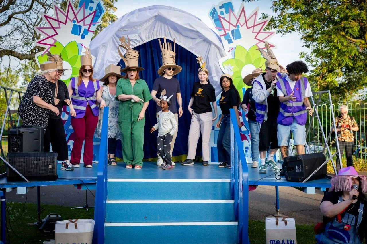 People performing a theatrical play on an outdoor stage, some wearing costumes including hats and masks, with a colorful backdrop and audience members nearby.