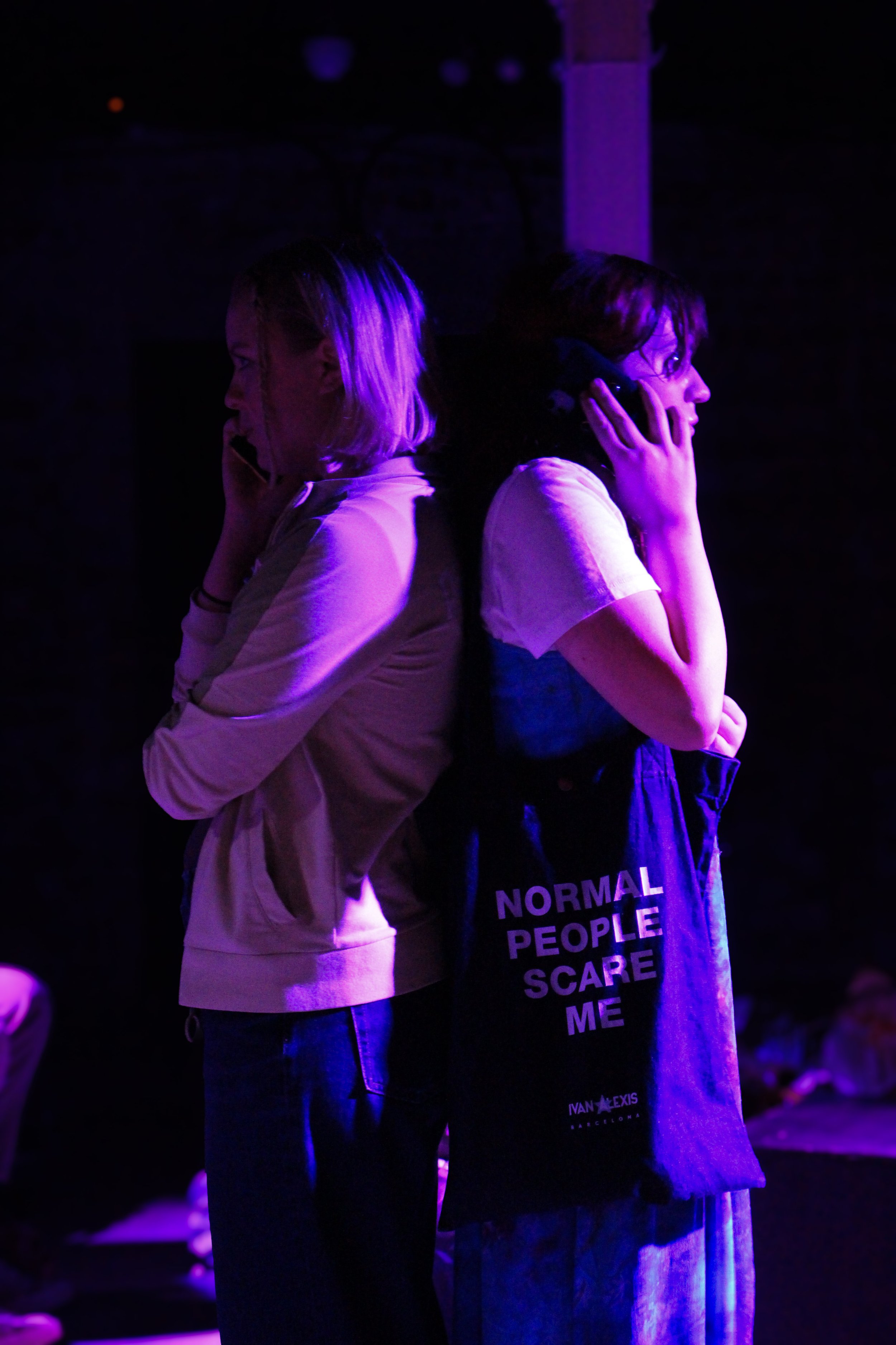 Two women are standing back-to-back under purple lighting, each holding a phone to their ear, one wearing a beige hoodie and the other a black shirt with white text that reads, 'NORMAL PEOPLE SCARE ME.'