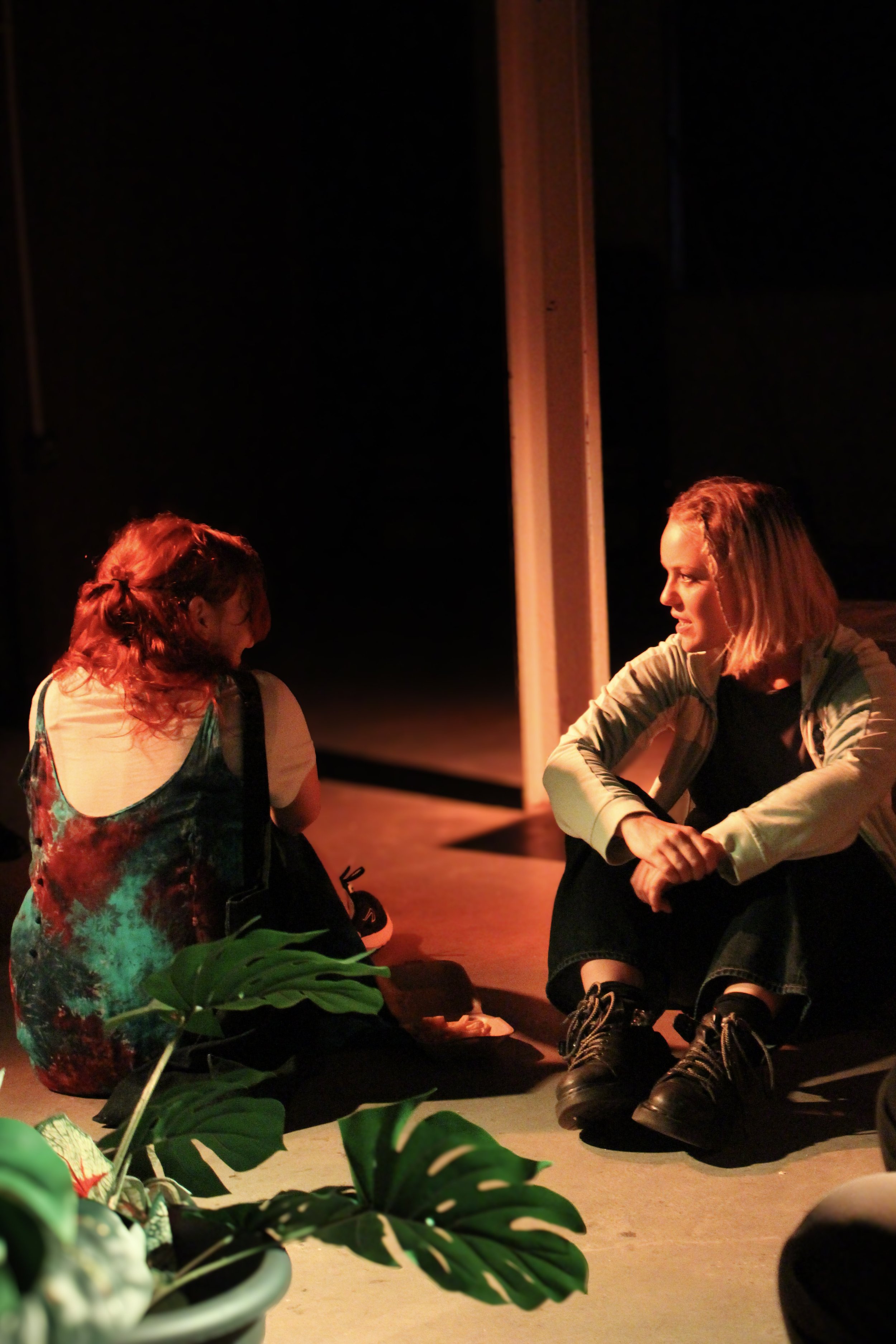 Two women sitting on the floor having a conversation in a dimly lit room. One woman has red hair and is wearing a colorful dress, while the other has blonde hair, braided, and is wearing dark clothing including boots. There is a large plant in the fo