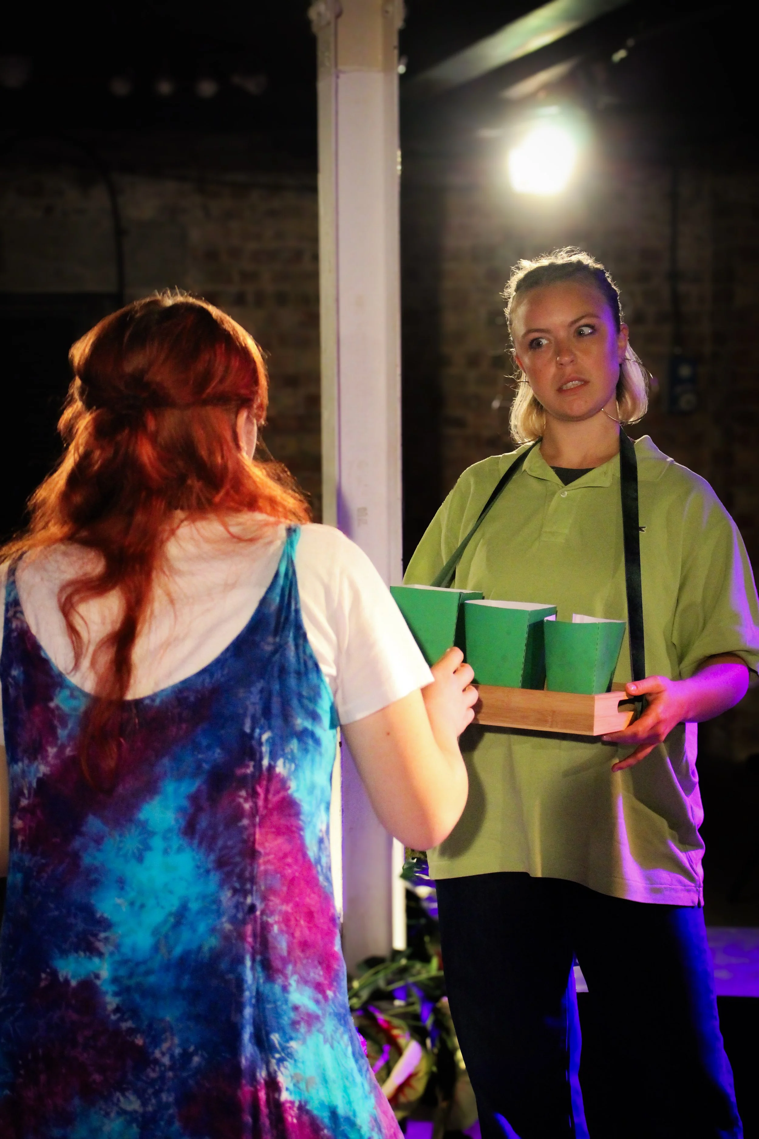 A woman in a green shirt is handing a tray with green containers to a girl with red hair and a colorful tie-dye shirt. The scene appears to be indoors with warm lighting and a brick wall in the background.