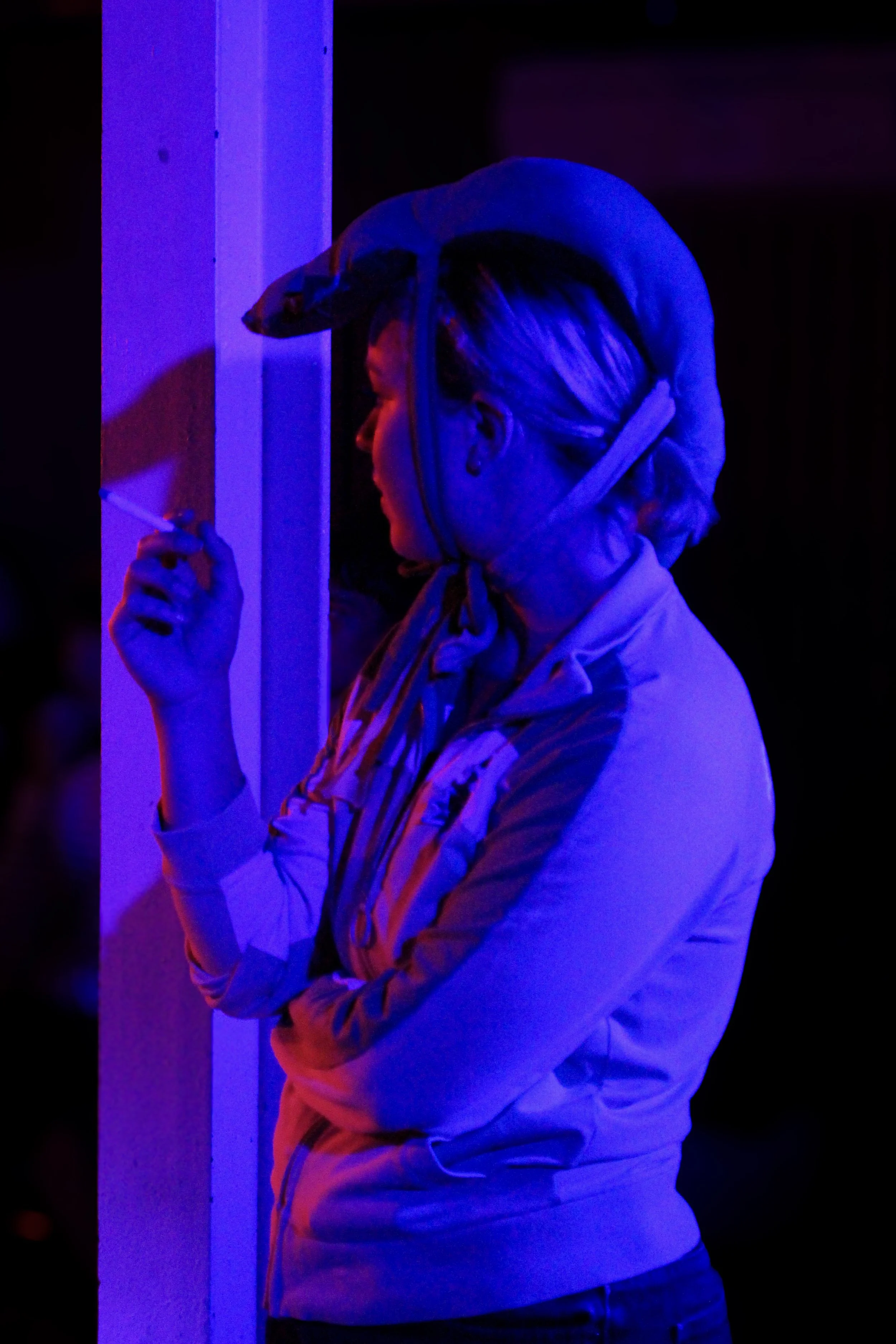 A woman with blonde hair styled in a messy bun, wearing a grey hoodie with red, yellow, and blue striped lining, stands in a dark room illuminated by blue and purple neon lights. She is holding a cigarette in her right hand while her gaze is directed