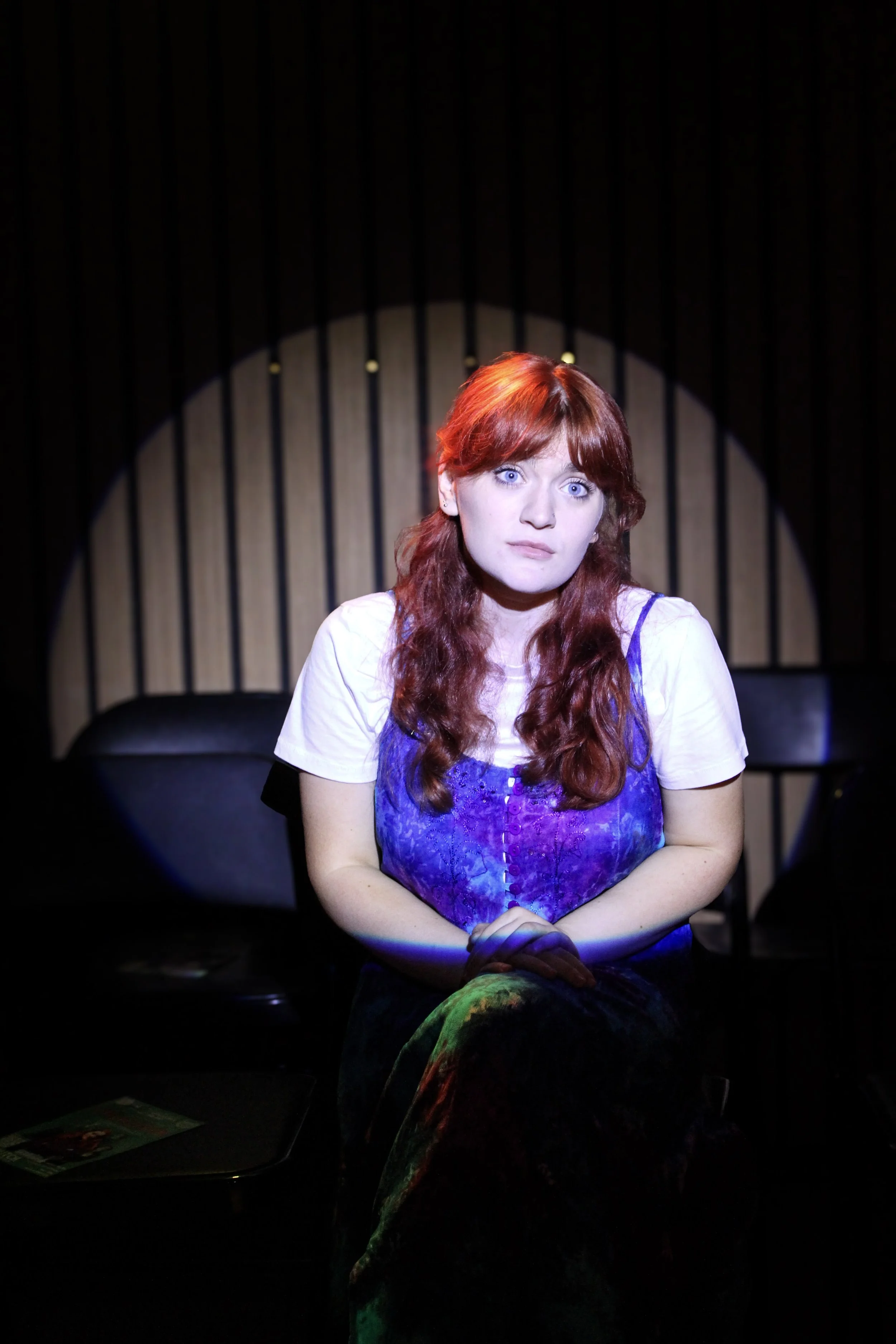 A woman with red hair and blue eyes sitting on a black chair, wearing a purple blouse and a white T-shirt, under a spotlight against a dark background with vertical bars.