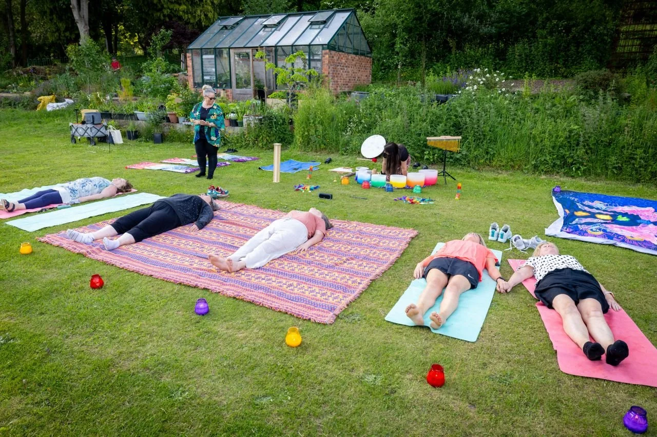 People participating in an outdoor yoga or meditation session on a grassy lawn, with some lying on mats and others standing nearby, surrounded by colorful candles and plants, with a greenhouse and trees in the background.