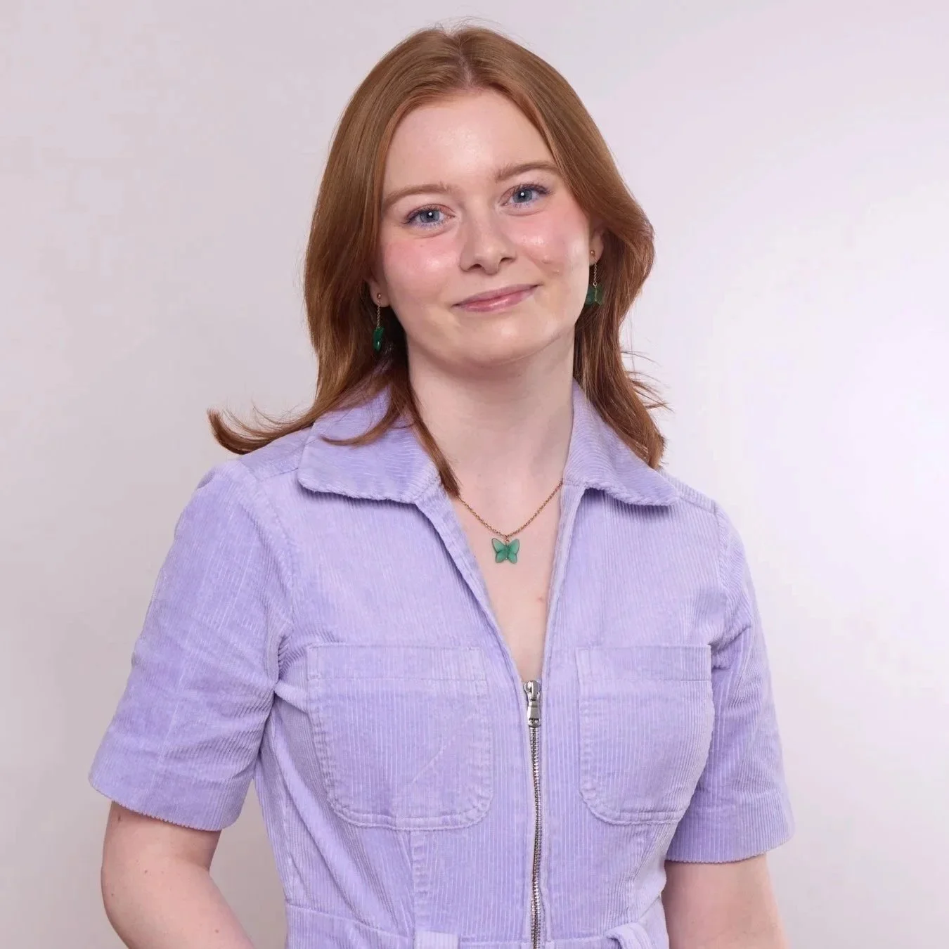 A woman with red hair and blue eyes wearing a lavender zip-up jacket, green butterfly jewelry, and a matching necklace, standing against a plain light-colored background.