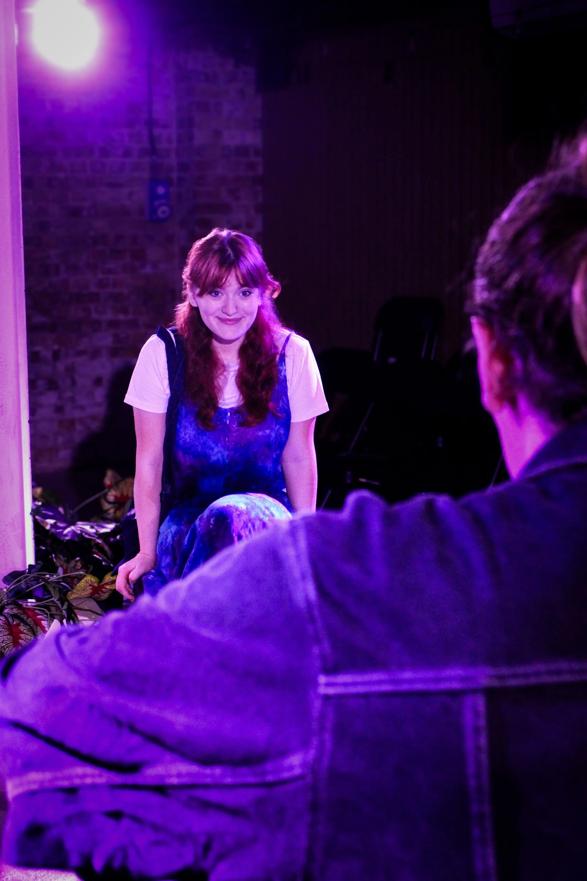 A woman with red hair and a joyful expression, wearing a white t-shirt and a blue dress, in a room illuminated with purple lighting, with a brick wall in the background, sitting on a chair.