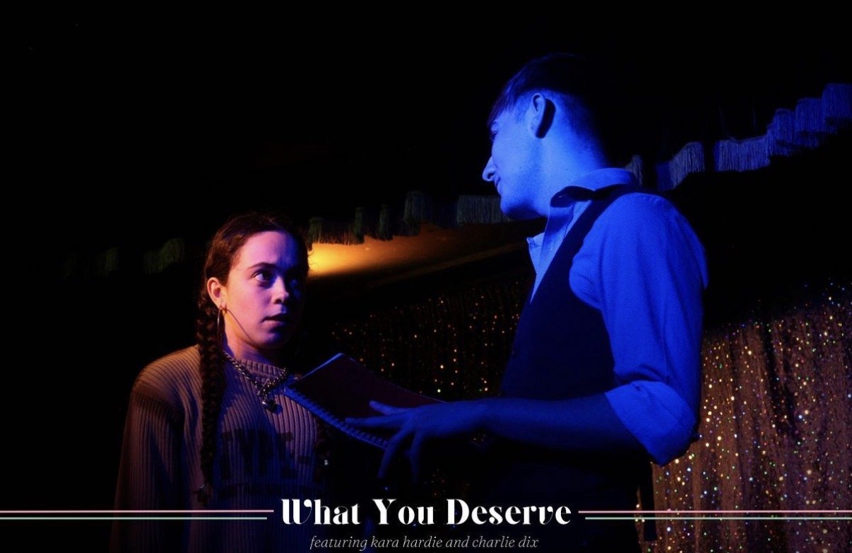 A young woman with braided hair and a young man holding a notebook, engaged in a serious conversation on a stage with a glittery backdrop, under dim blue and orange lighting.