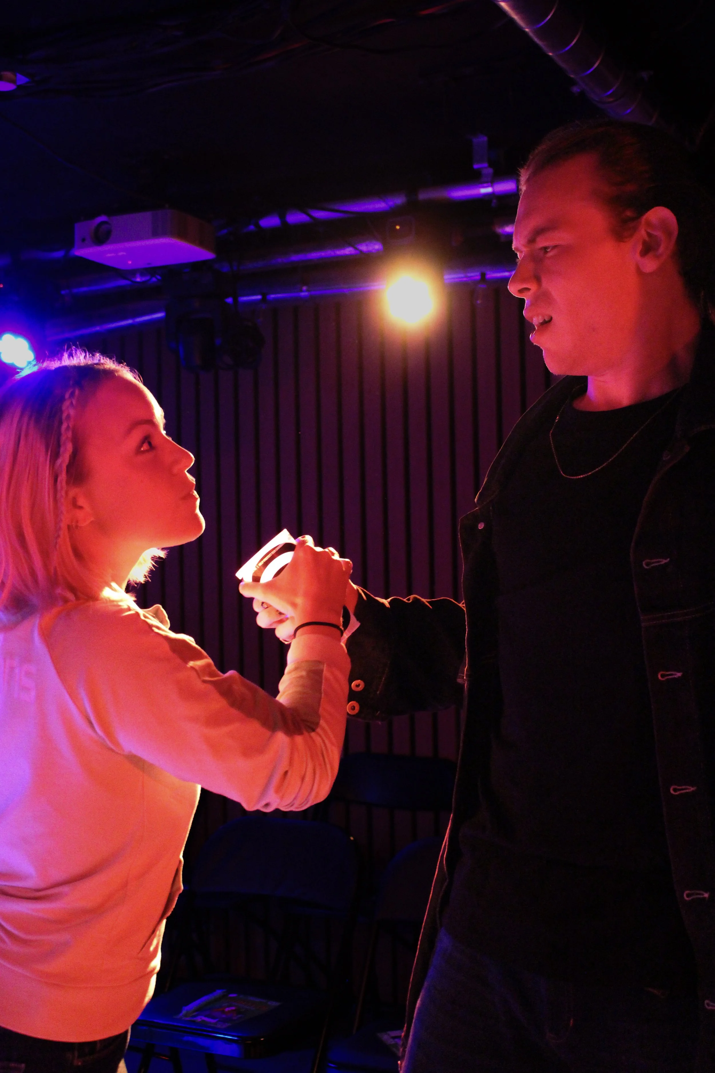 A young woman with blonde hair and a young man with dark hair engaging in a conversation in a dimly lit room with purple and yellow stage lighting.