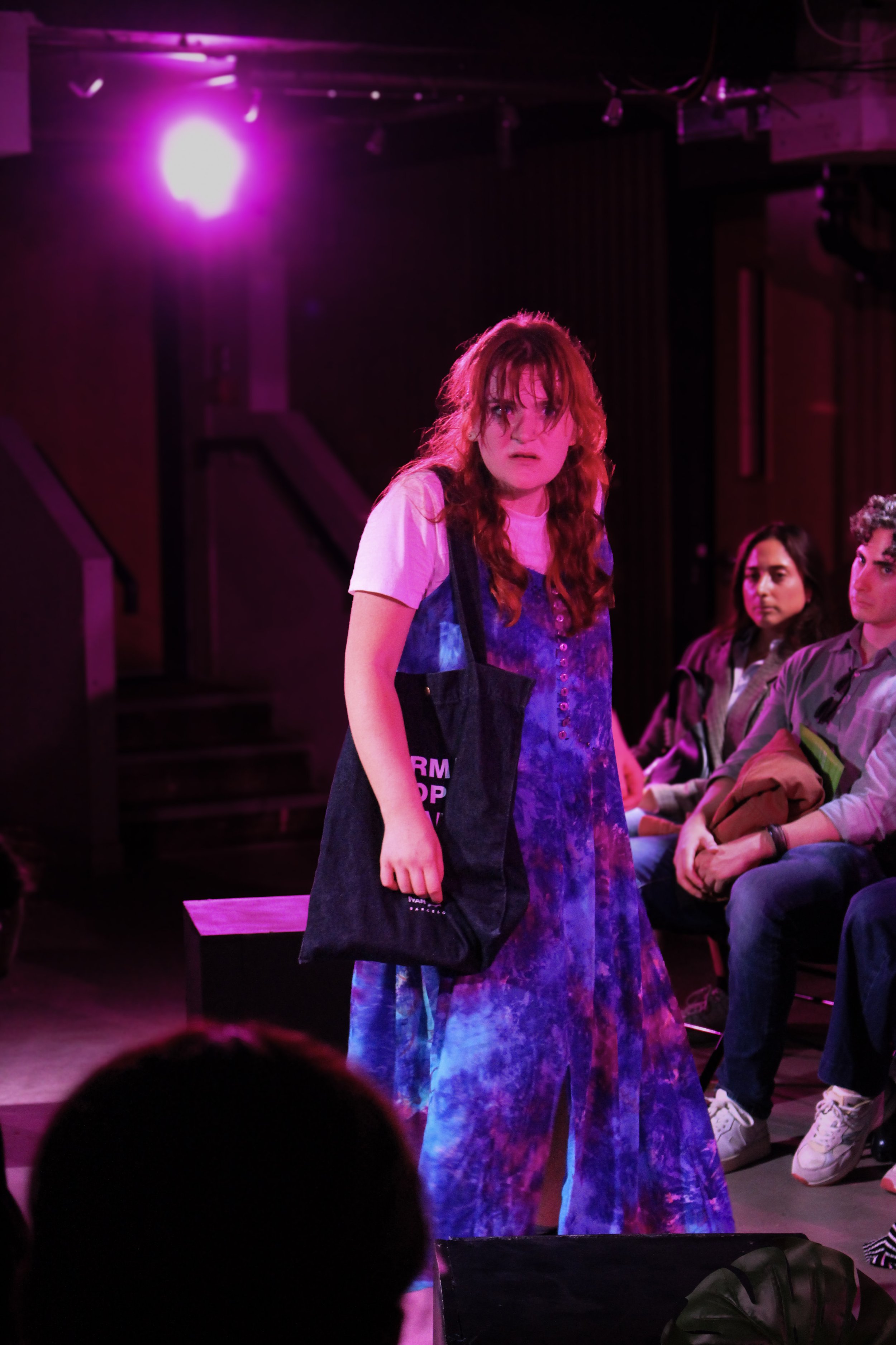 Young woman with long red hair, wearing a purple tie-dye dress and jean jacket, standing under purple stage lighting with a dark background, and other seated people visible in the background.