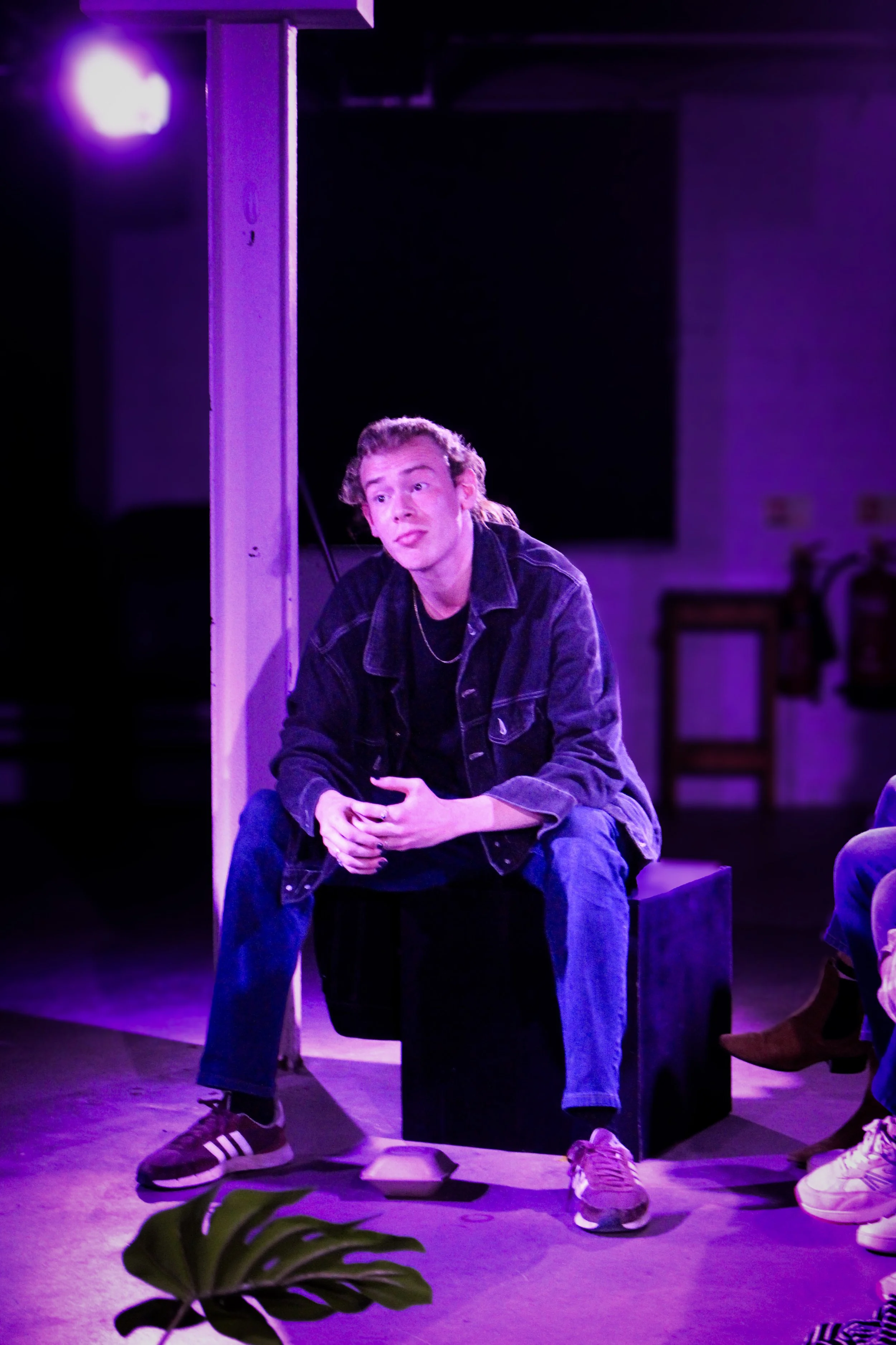 A young man with curly hair sitting on a black box, wearing a denim jacket and jeans, under purple stage lighting in a room with dark walls and chairs.
