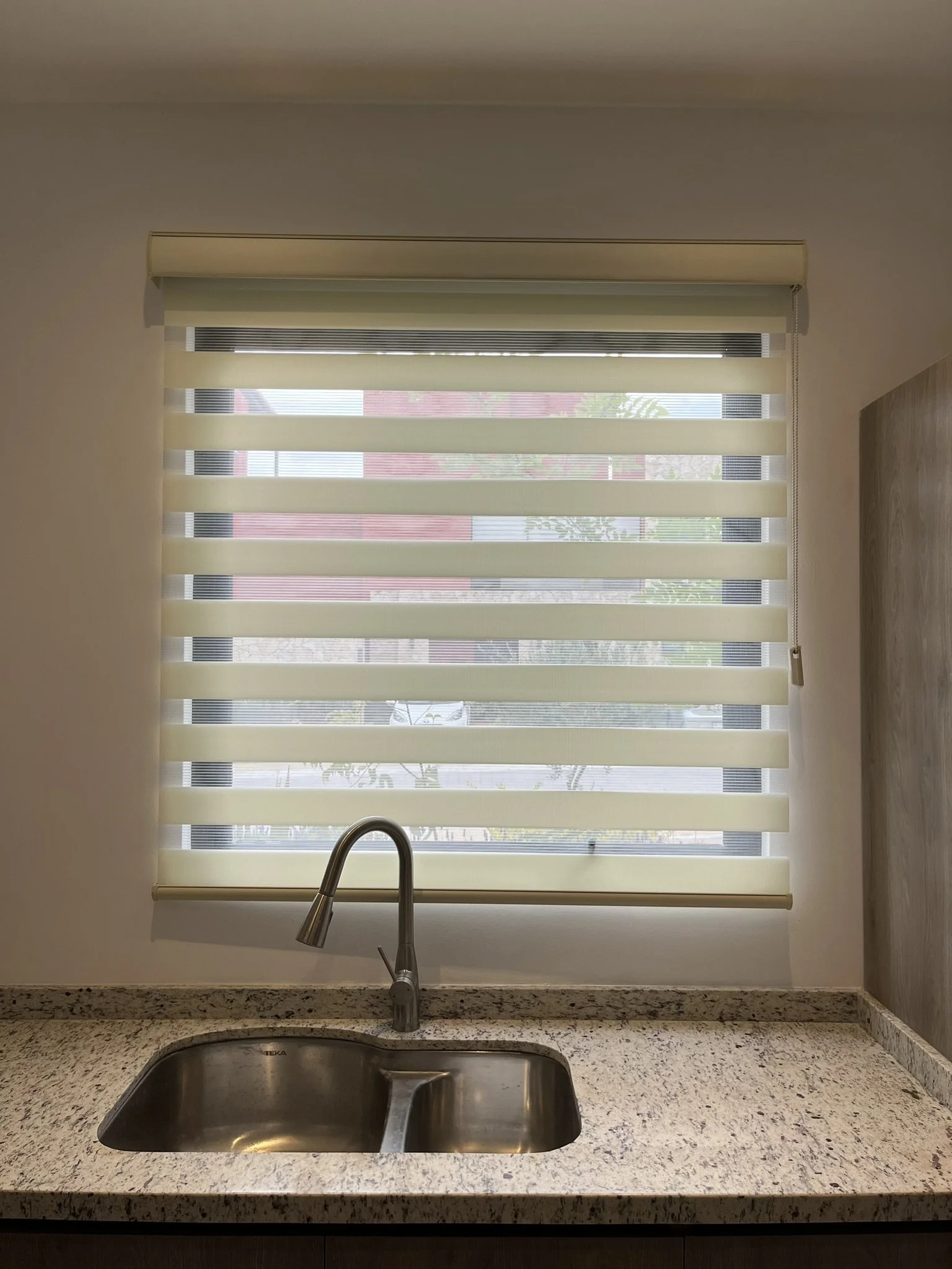Vista de una cocina con fregadero de acero inoxidable, superficie de granito gris y una ventana con persianas sheer elegance color blanco. Apartamento en San Miguel de Allende