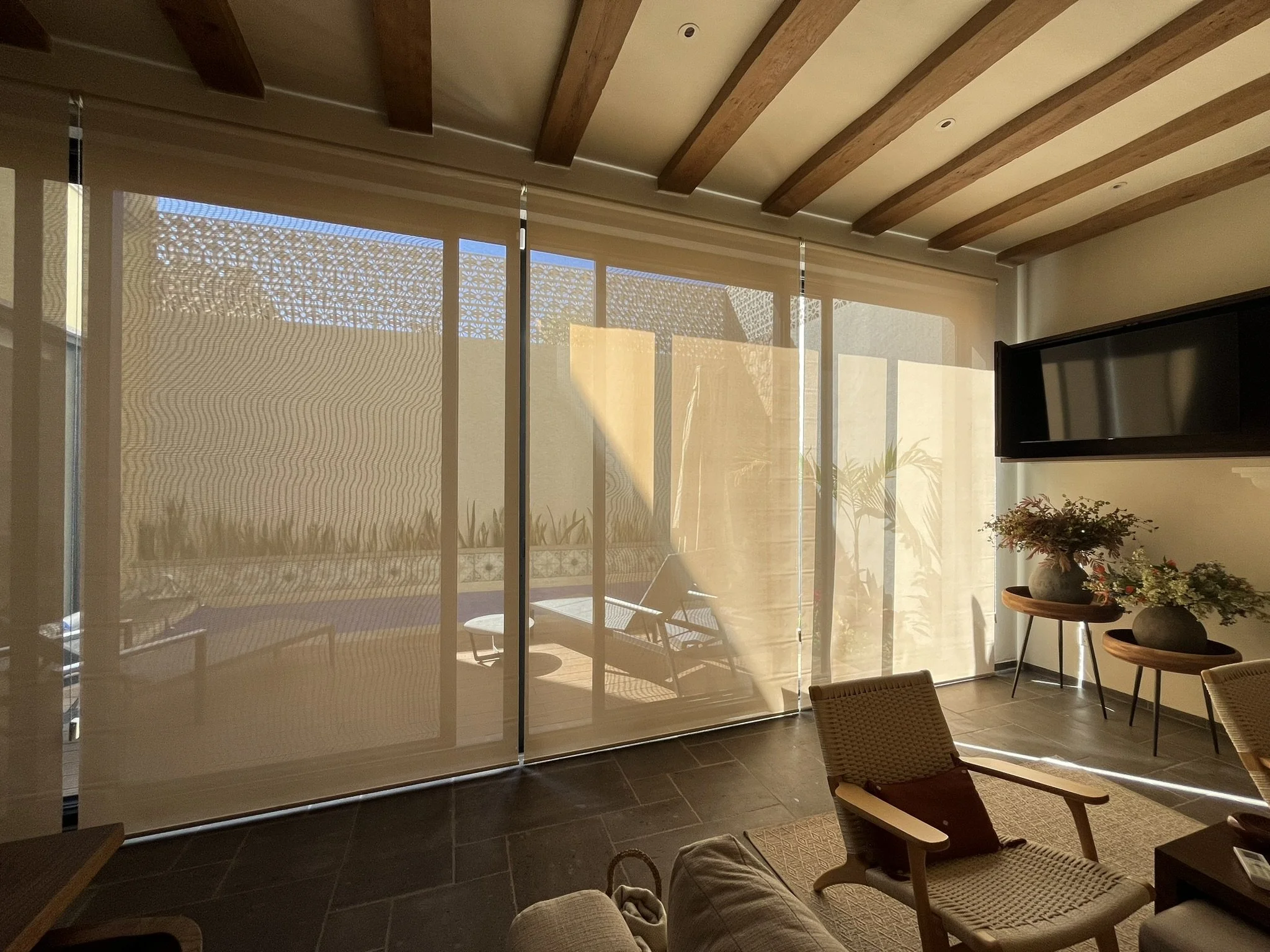 Interiores de sala con paredes de cristal, flores en dos macetas, y sillas de madera y mimbre, con sombra del exterior en la cortina y el suelo de piedra. Ventanas con persianas enrollables traslúcidas o de malla solar.