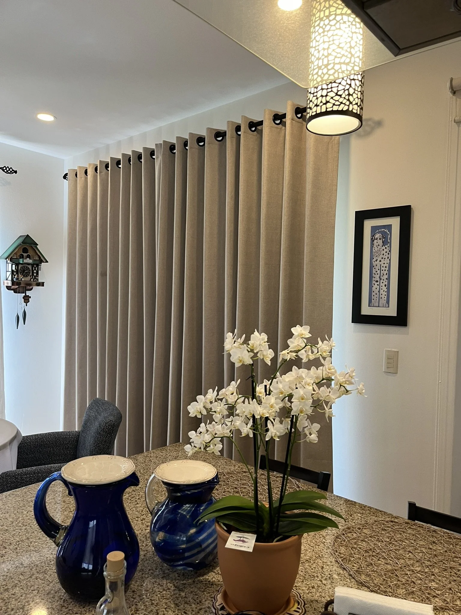 Vase con orquídeas blancas en una mesa de comedor, con platos y jarras azules, en una sala decorada con cortinas beige de estilo ojillo u ojal, en un cortinero redondo de madera color caoba, un reloj de cuco y una pintura en la pared.