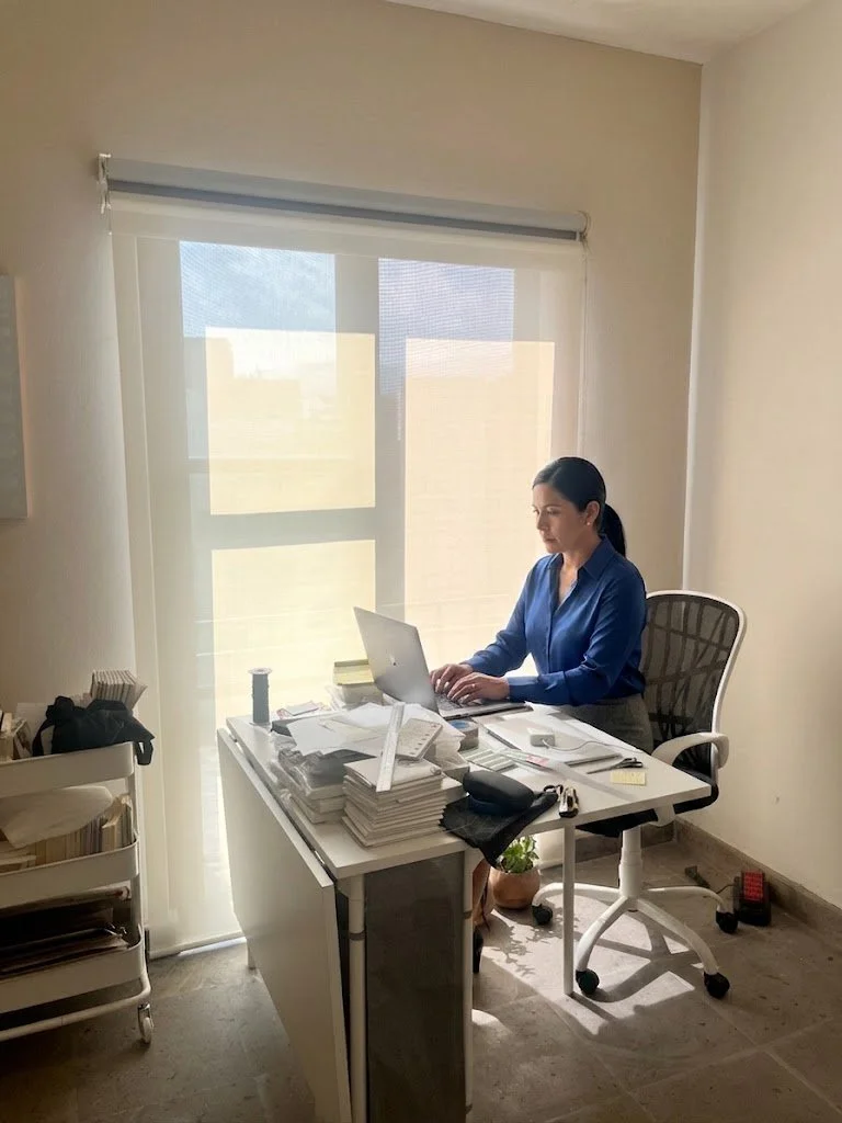 Mujer trabajando en una oficina con un escritorio lleno de papeles, laptops y otros objetos. La mujer está sentada frente a una ventana cubierta con una cortina translúcida, usando una blusa azul.