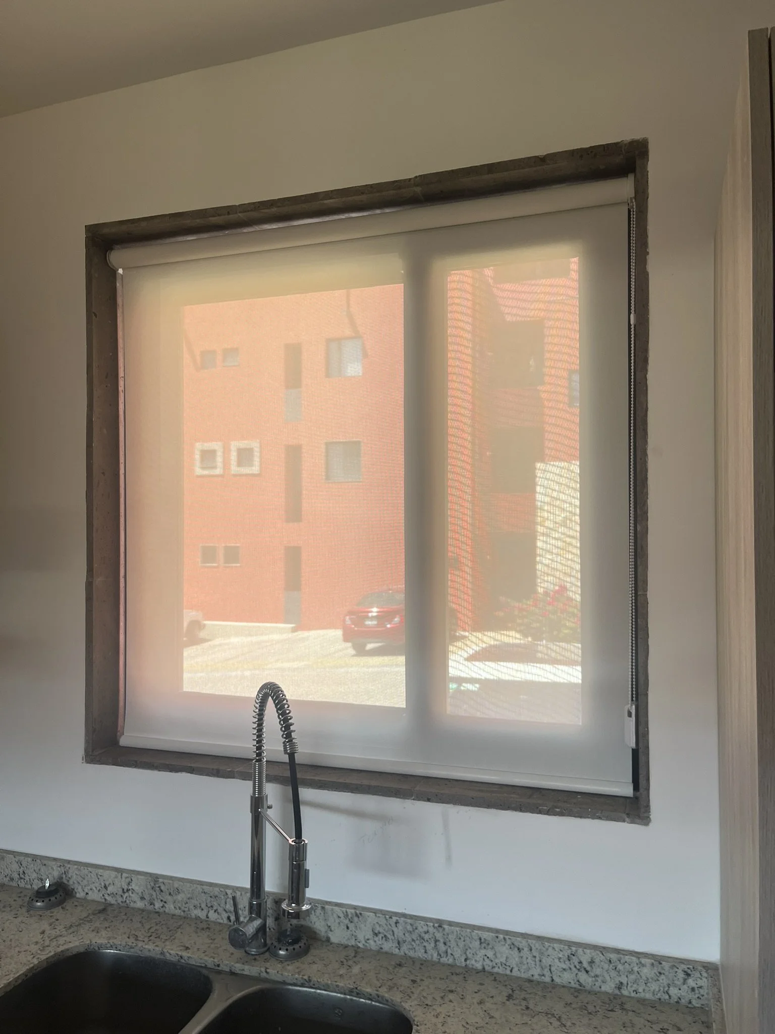 Ventana con reja y persiana enrollable traslúcida en una oficina o cocina, vista del exterior con edificios y un auto. Está instalada sobre una mesada de granito con fregadero y manguera de cocina.