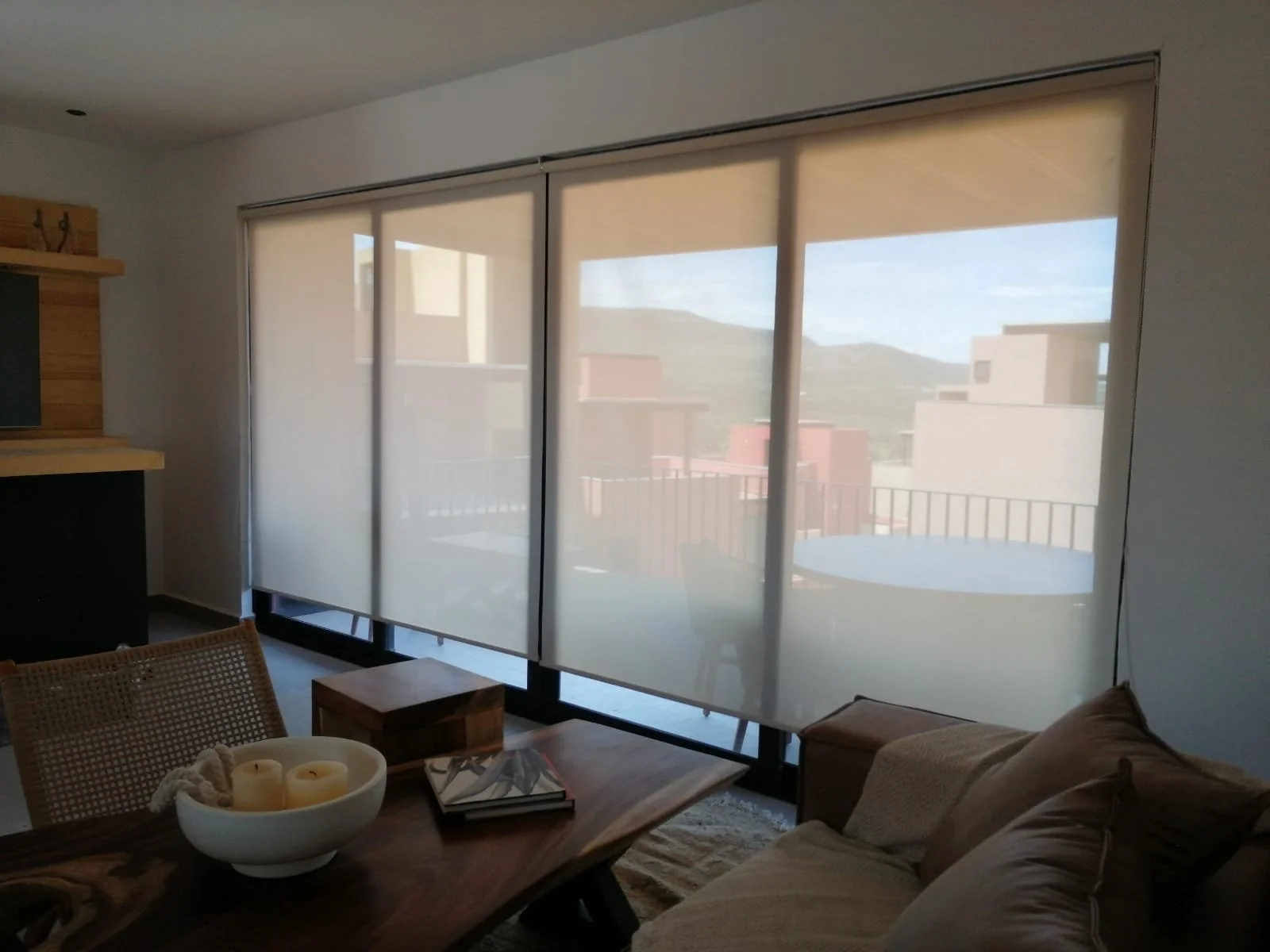 Sala de estar con ventanas de persianas enrollables blancas tipo malla solar, sillón beige, mesa de madera, decoraciones y vista panorámica de edificios y montañas. Fraccionamiento en San Miguel de Allende.