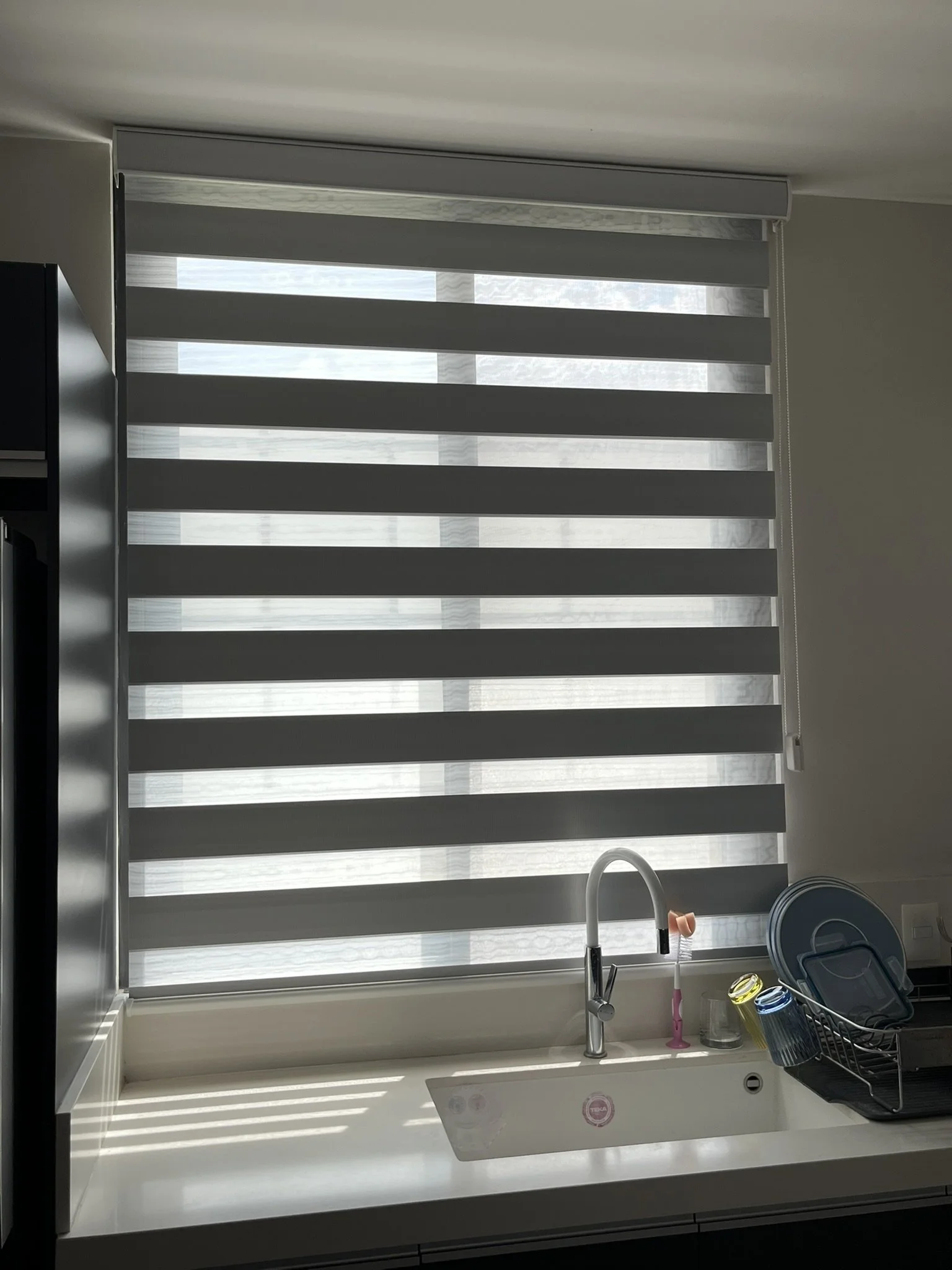 Vista de una cocina con ventana con persiana Sheer Elegance en tonos grises, fregadero con grifo, un dispensador de jabón rosa, y utensilios de limpieza en un escurridor negro junto a la piedra de la encimera. Apartamento en San Miguel de Allende