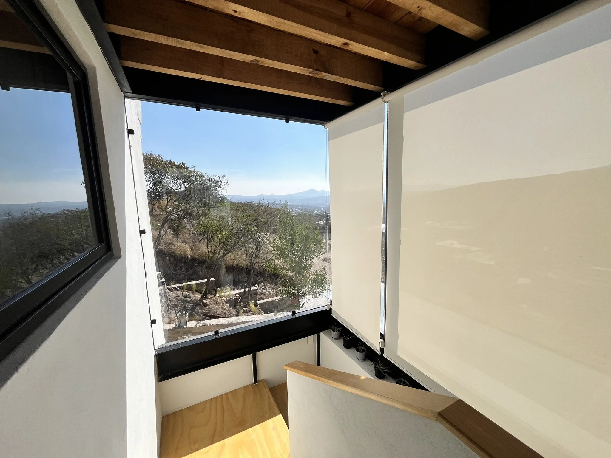 Vista desde un balcón o ventana interior que muestra una escena exterior con árboles y montañas en el horizonte, en un día despejado. Ventana con persianas enrollables traslúcidas que protegen del sol