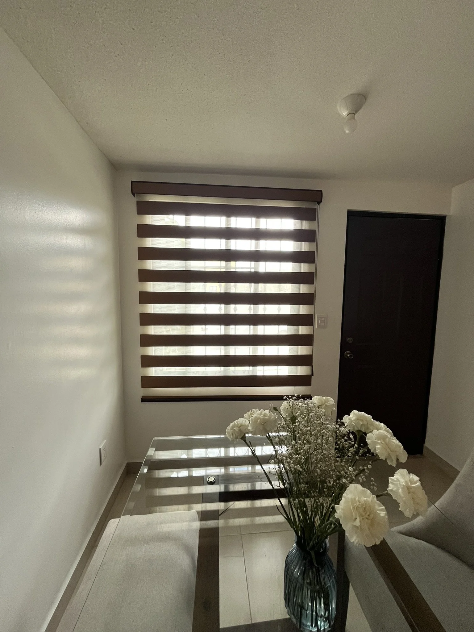 Interior de una sala con una mesa de cristal, florero con flores blancas y cortinas sheer elegance en la ventana