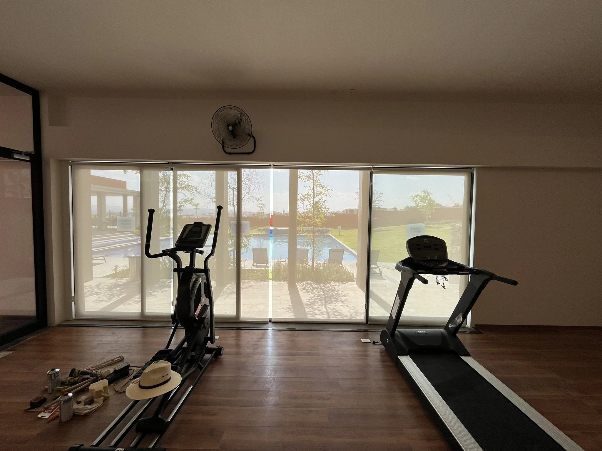 Gimnasio con máquinas, vista a través de ventanas de un exterior con piscina y árboles. Las ventanas tiene persianas enrollables tipo malla solar o traslúcidas. Fraccionamiento en San Miguel de Allende.