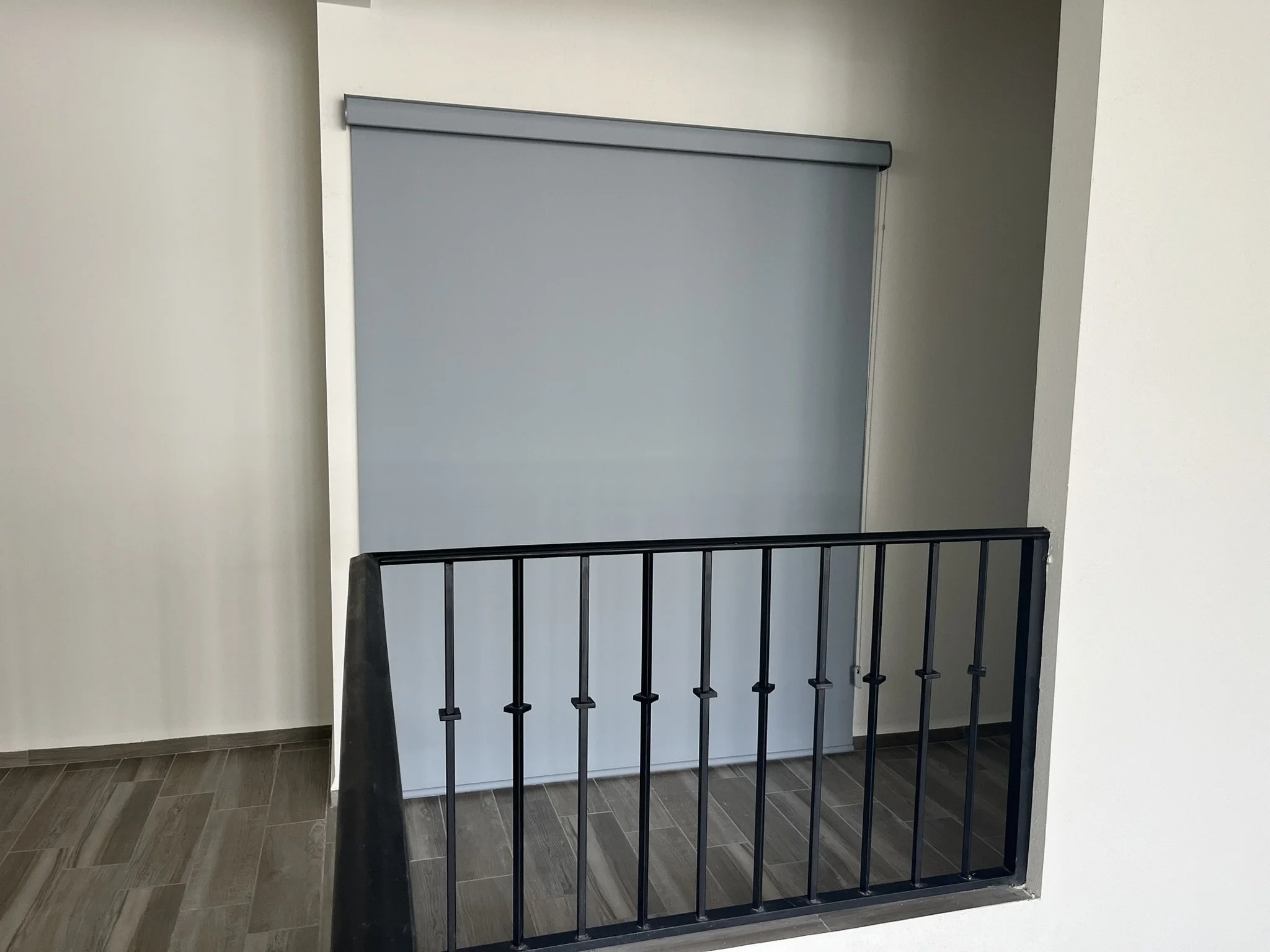 Barandal de pasamanos negro con paneles de vidrio y un muro gris en el fondo, en un pasillo del interior de un edificio.