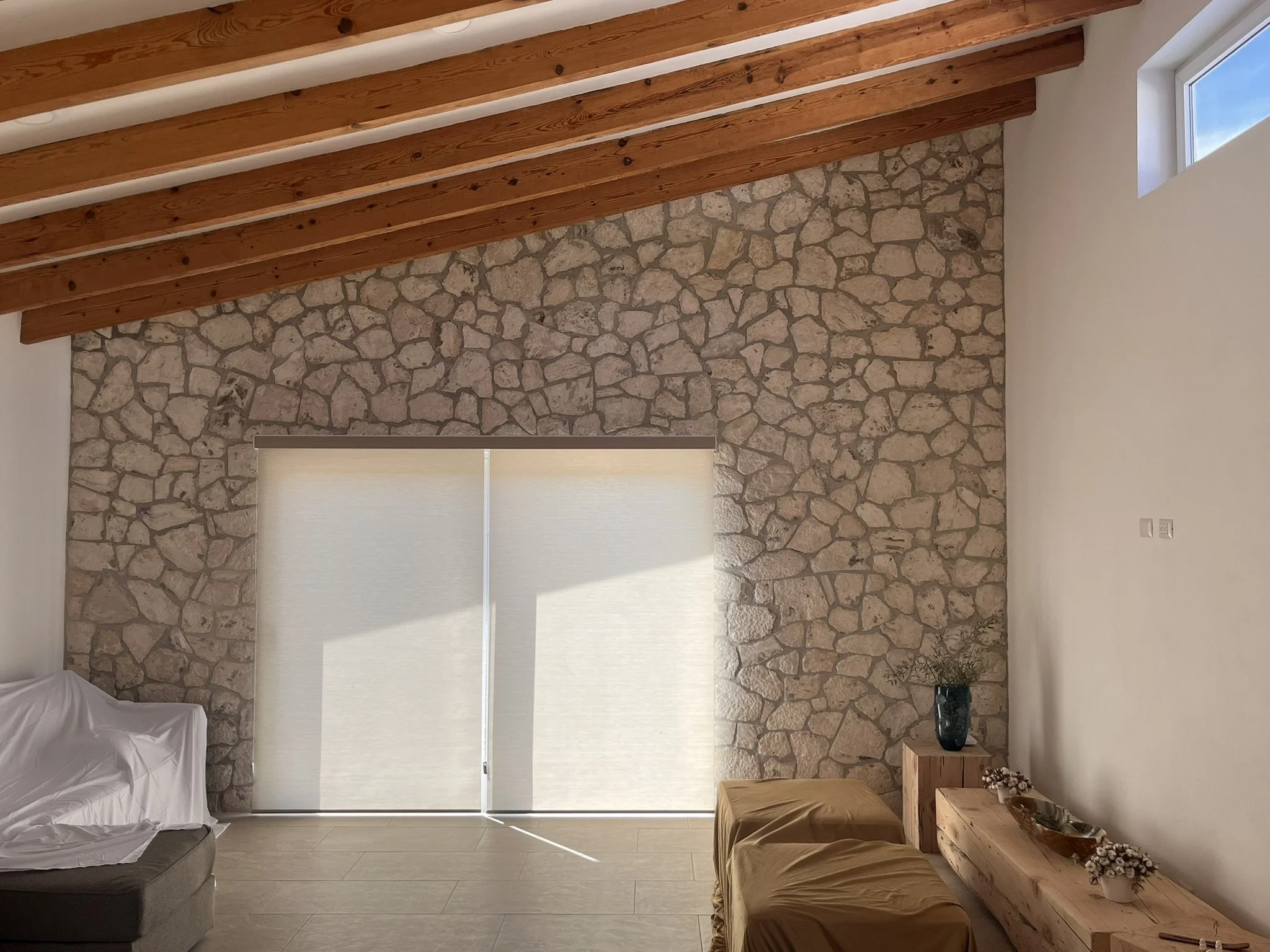 Sala con pared de piedra y techo de vigas de madera, con ventanas con persianas enrollables de estilo semi-traslúcidas, un sofá y una mesa de madera con decoraciones.