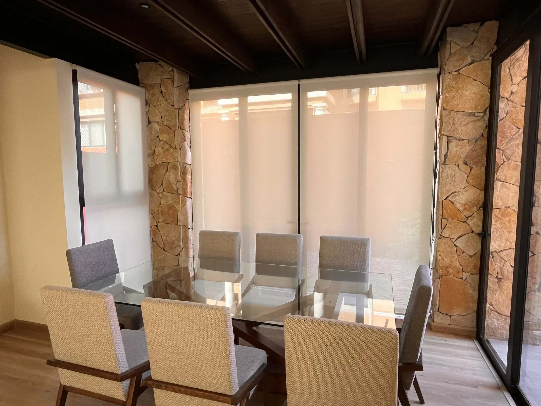Comedor con mesa de vidrio y sillas beige, paredes de piedra y ventanas con persianas enrollables blancas. Fraccionamiento en San Miguel de Allende.