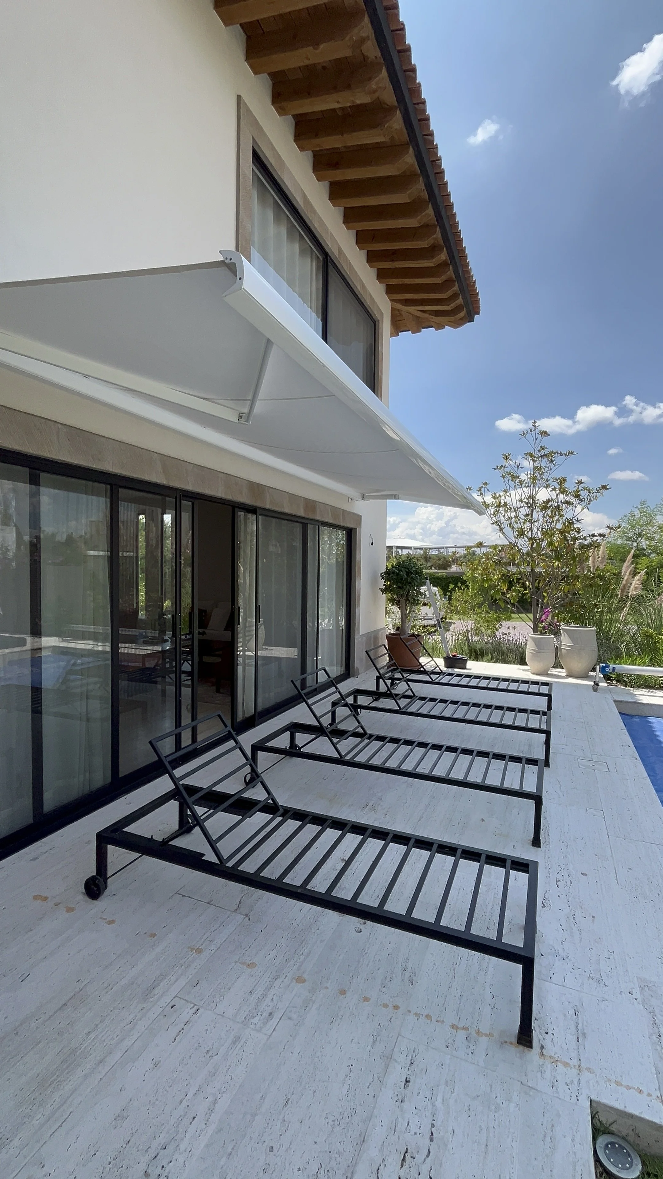 Toldo retráctil o de proyección, color blanco, tipo cassette, en una Terraza exterior con sillas de diseño negro, macetas grandes con plantas, y una alberca, vista de una casa moderna con ventanas grandes y un toldo blanco, bajo un cielo azul.