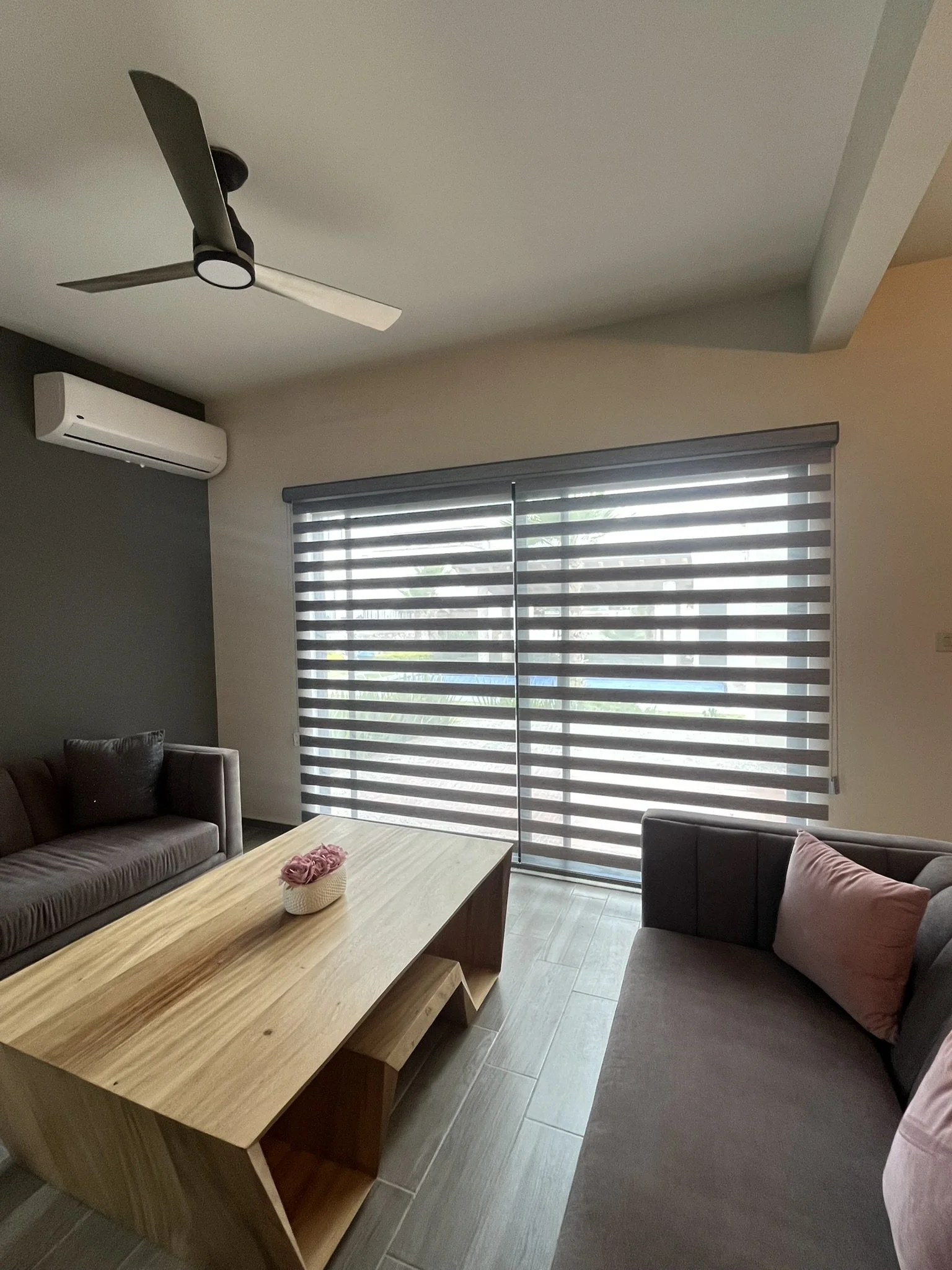 Sala de estar con sillones gris oscuro, mesa de madera en el centro, cortinas enrollables estilo sheer elegance en la ventana y un aire acondicionado. Casa en San Miguel de Allende.