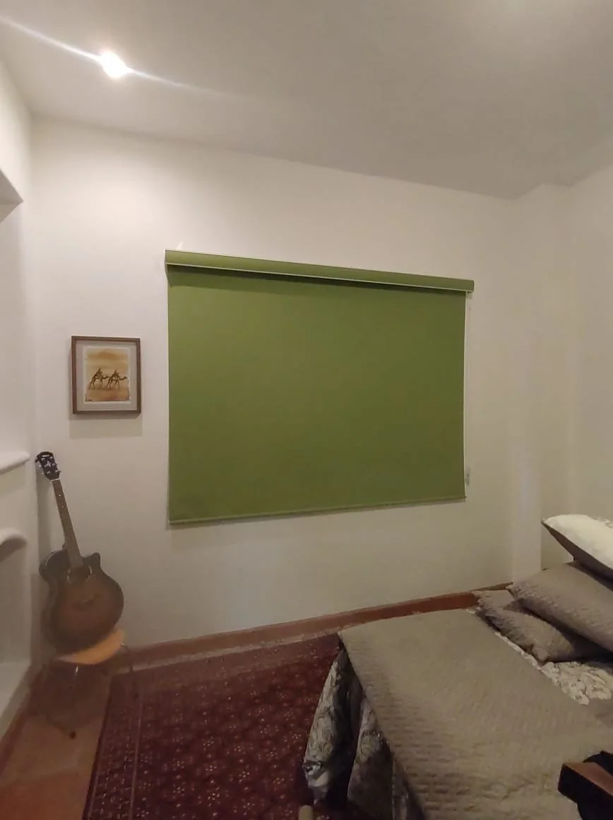 Habitación con pared blanca, ventana con cortina verde, cuadro de caballos, guitarra apoyada en una pequeña silla, cama con sábanas y almohadas, alfombra en el suelo.