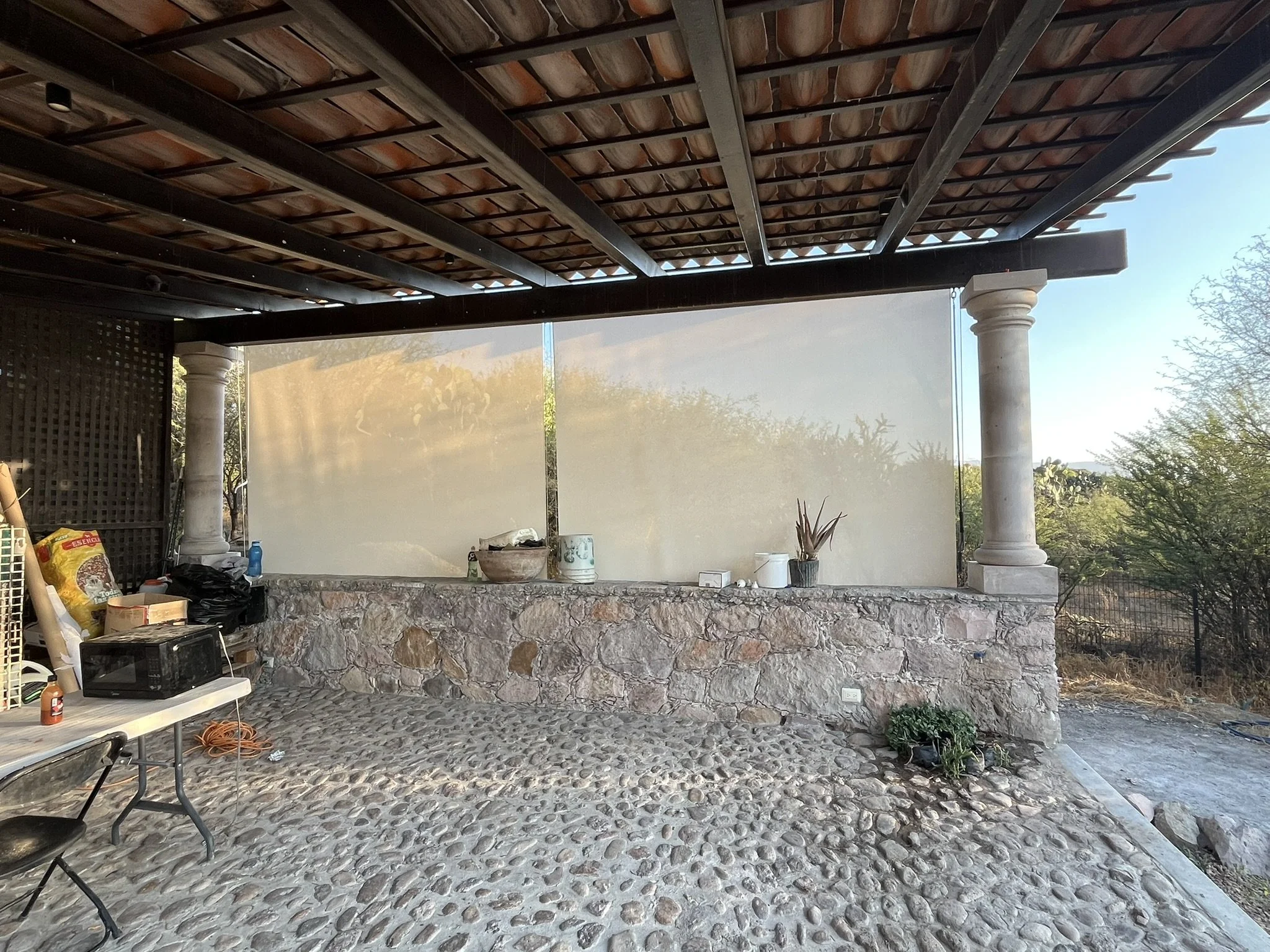 Una estructura de pérgola de madera con columnas de piedra decorativa, techo de tejas y un área de patio con piso de piedras, con objetos y plantas dispersas a su alrededor. Al fondo, dos cortinas enrollables para protección solar y del viento.
