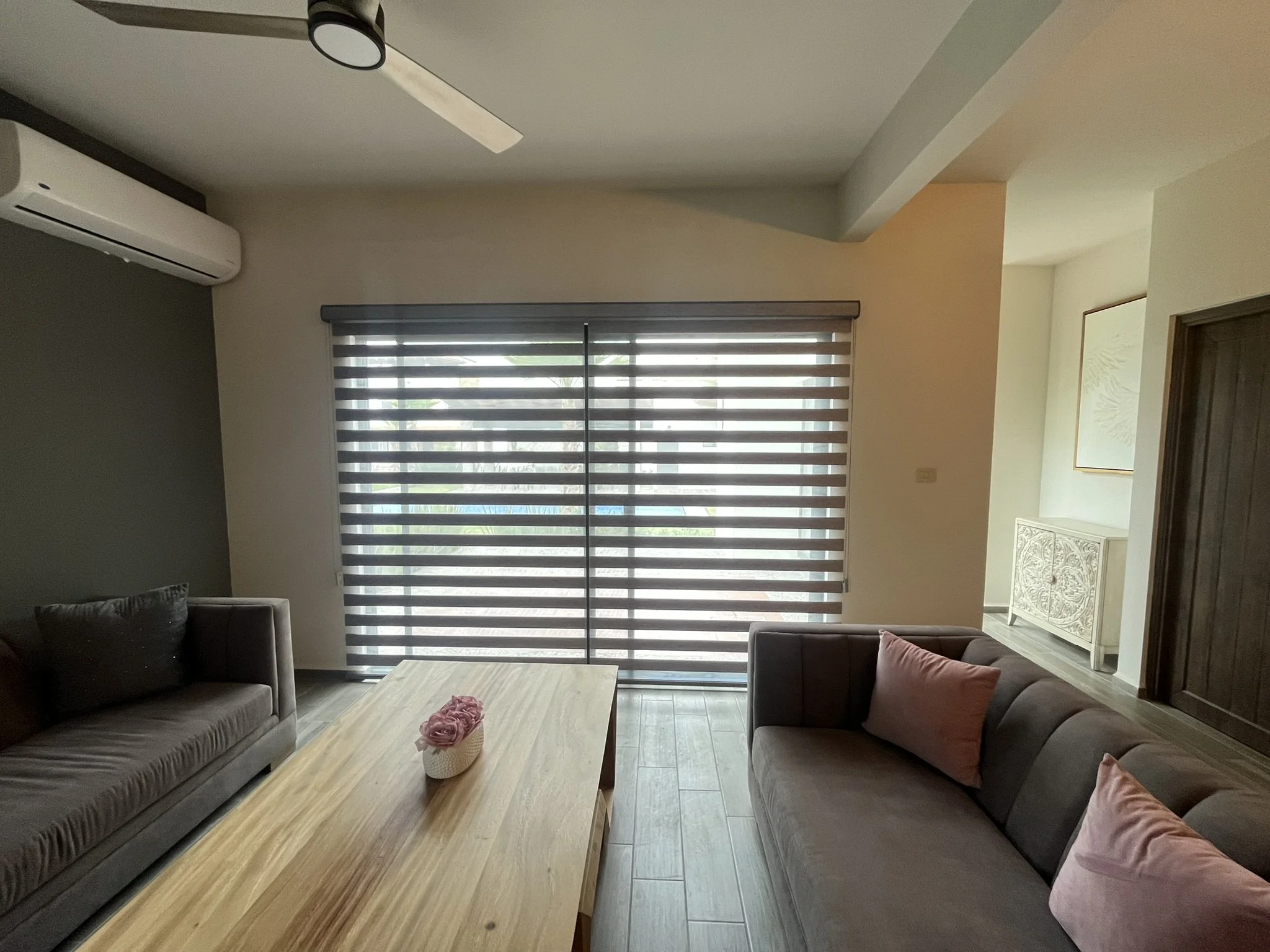 Sala de estar con tres sillones; uno en color gris y los otros en tonos marrón y rosa, mesa de madera clara con adorno de flores en un florero, cortinas horizontales o Sheer Elegance en ventana grande que deja entrar luz natural, 