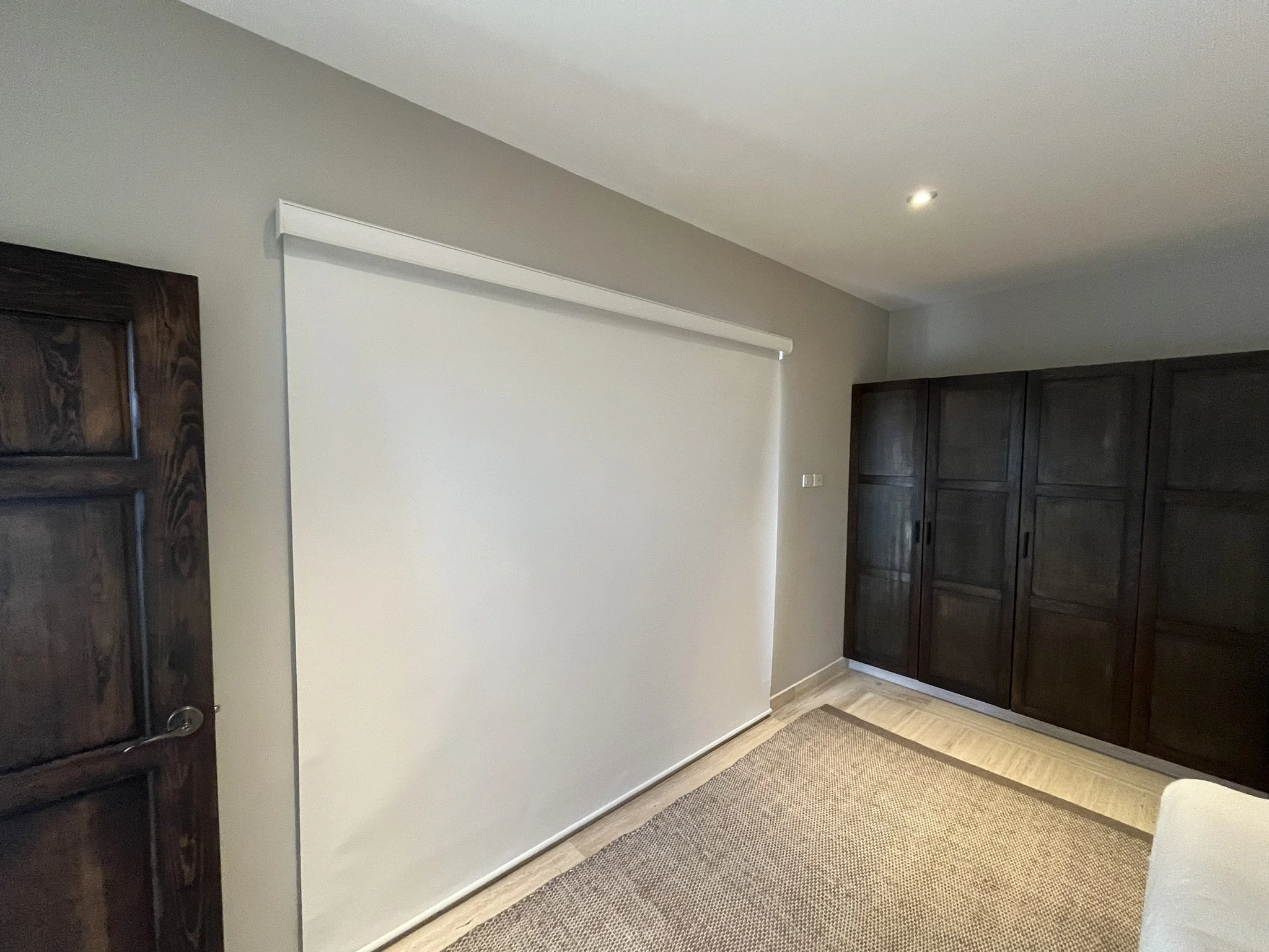 Sala de estar con pared blanca, una persiana blackout cubre la ventana. A la izquierda, el acceso a la recámara, una puerta de madera oscura y armario grande de madera oscura. Casa ubicada en fraccionamiento de San Miguel de Allende.