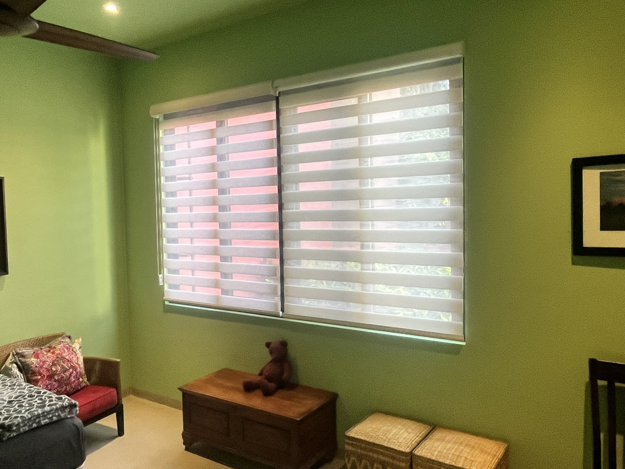 Una habitación con paredes verdes y ventana con cortinas estilo Sheer Elegance, de trama blanca y malla solar. Hay una cama con cobertor de diseño en la esquina izquierda, una mesa de madera con un oso de peluche, y varias sillas de mimbre apiladas.