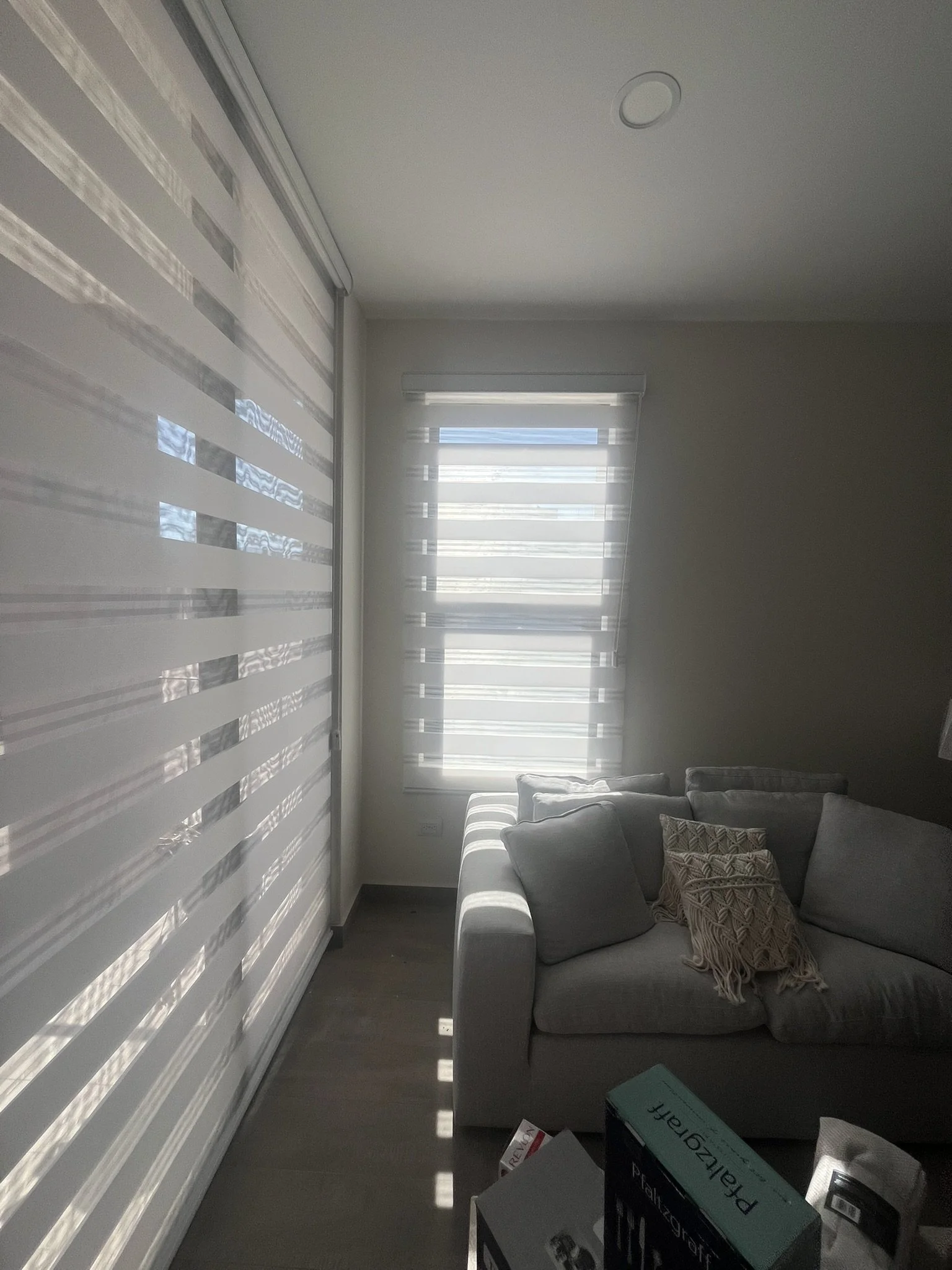 Sala con sofá gris, varias almohadas decorativas, ventana con persianas Sheer Elegance blancas, paredes blancas y suelo de madera. Departamento en San Miguel de Allende