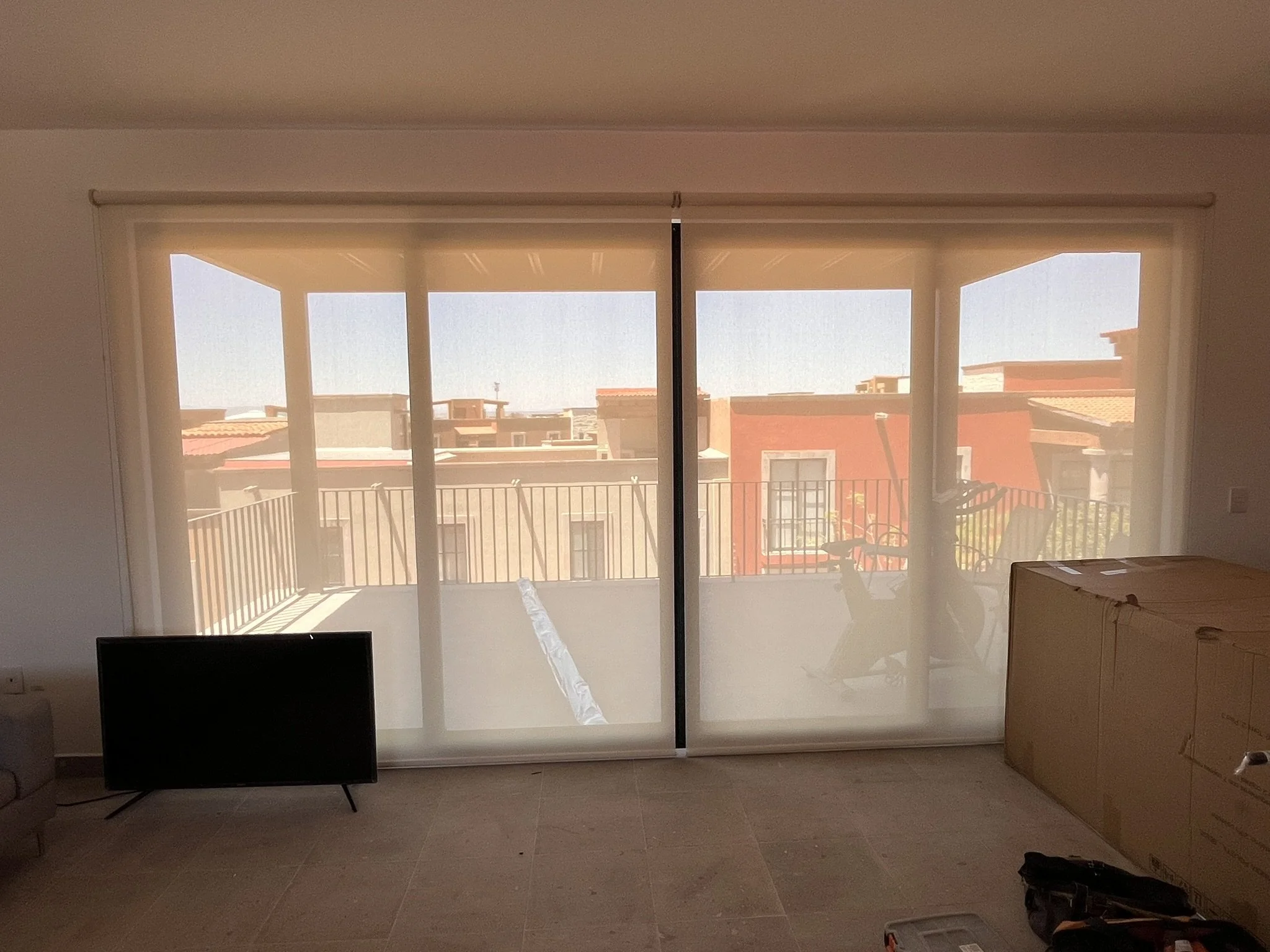 Habitación con piso de baldosas, un monitor en el suelo, caja de cartón y una ventana grande con cortinas enrollables traslúcidas que protegen del sol. Fuera de la ventana se ven edificios y un cielo despejado. Fraccionamiento en San Miguel de Allend