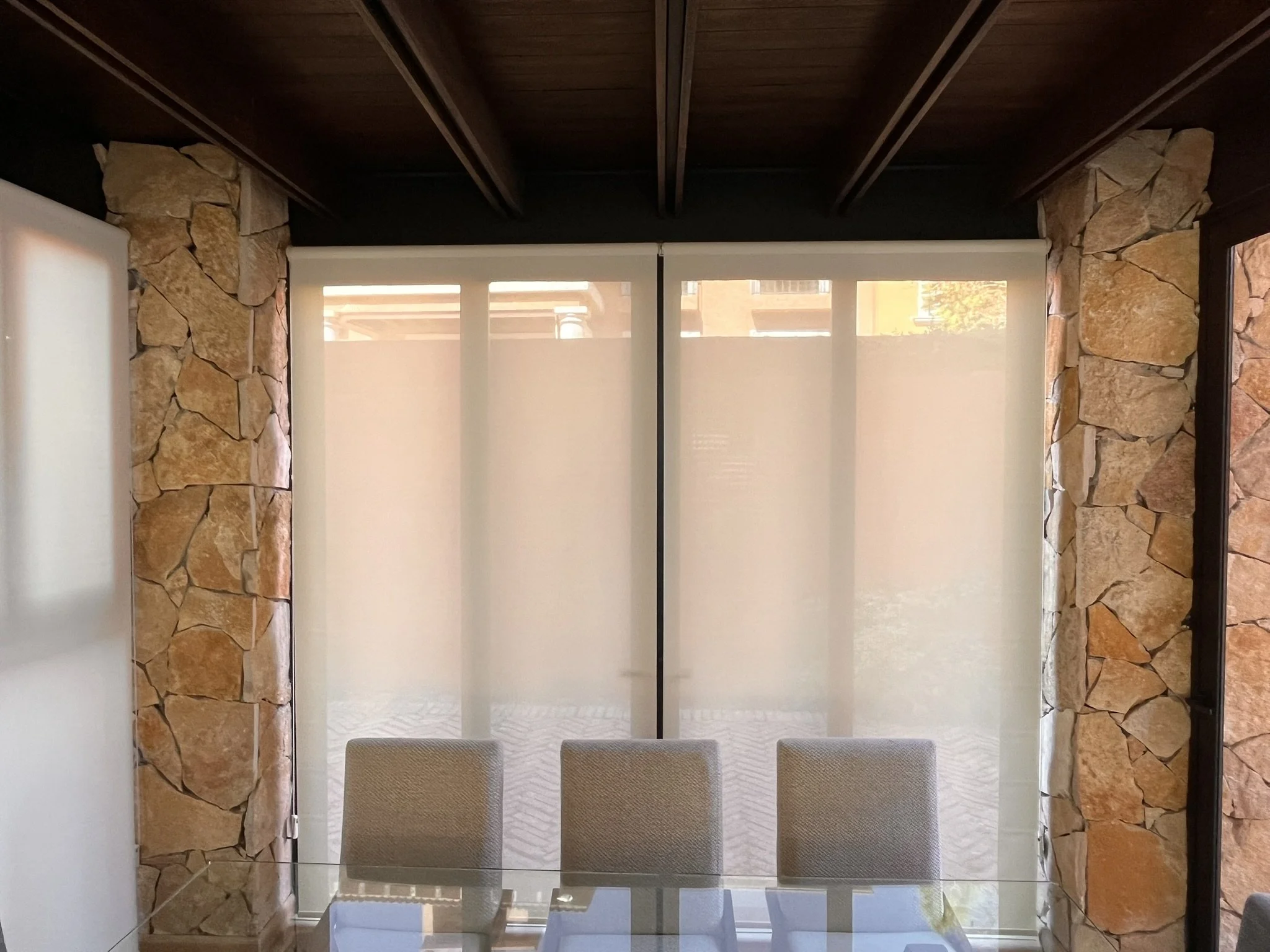 Vista de una sala con paredes de piedra y ventanas con persianas enrollables traslúcidas color blanco, con sillas de tela beige y una mesa de cristal.