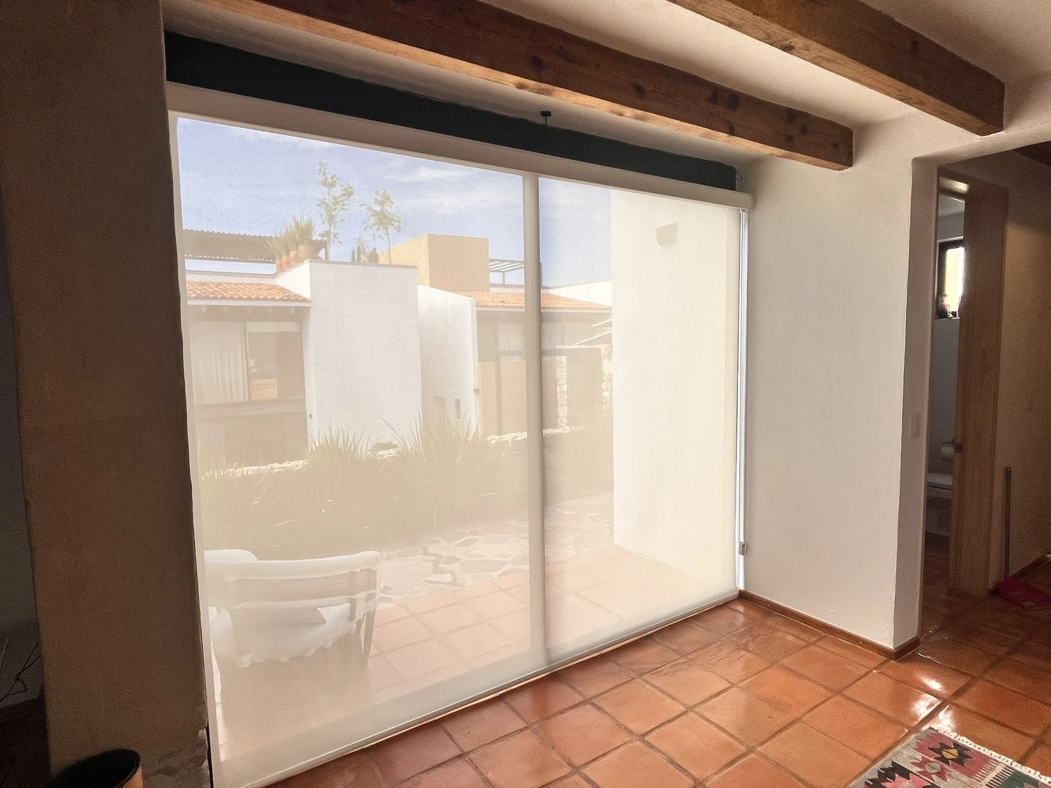 Ventana grande con cortina translúcida en un interior residencial, vista a casas con techos de tejas y árbol.