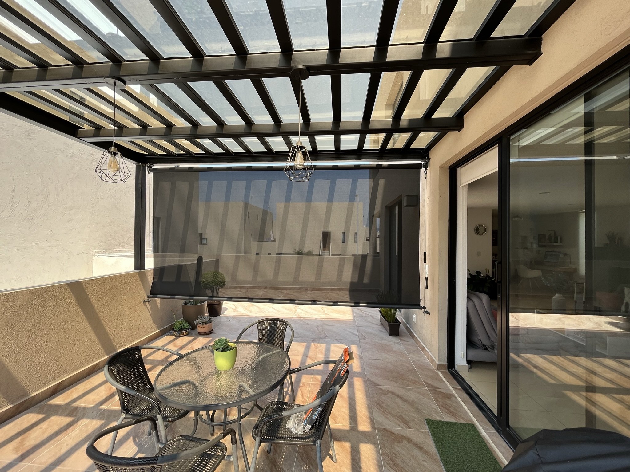 Al fondo, una cortina enrollable con tela tipo malla solar color negro. Terraza cubierta con pilares de metal, mobiliario de mimbre y una mesa de cristal con plantas decorativas, ventanas grandes de vidrio y sombra de estructuras de metal.
