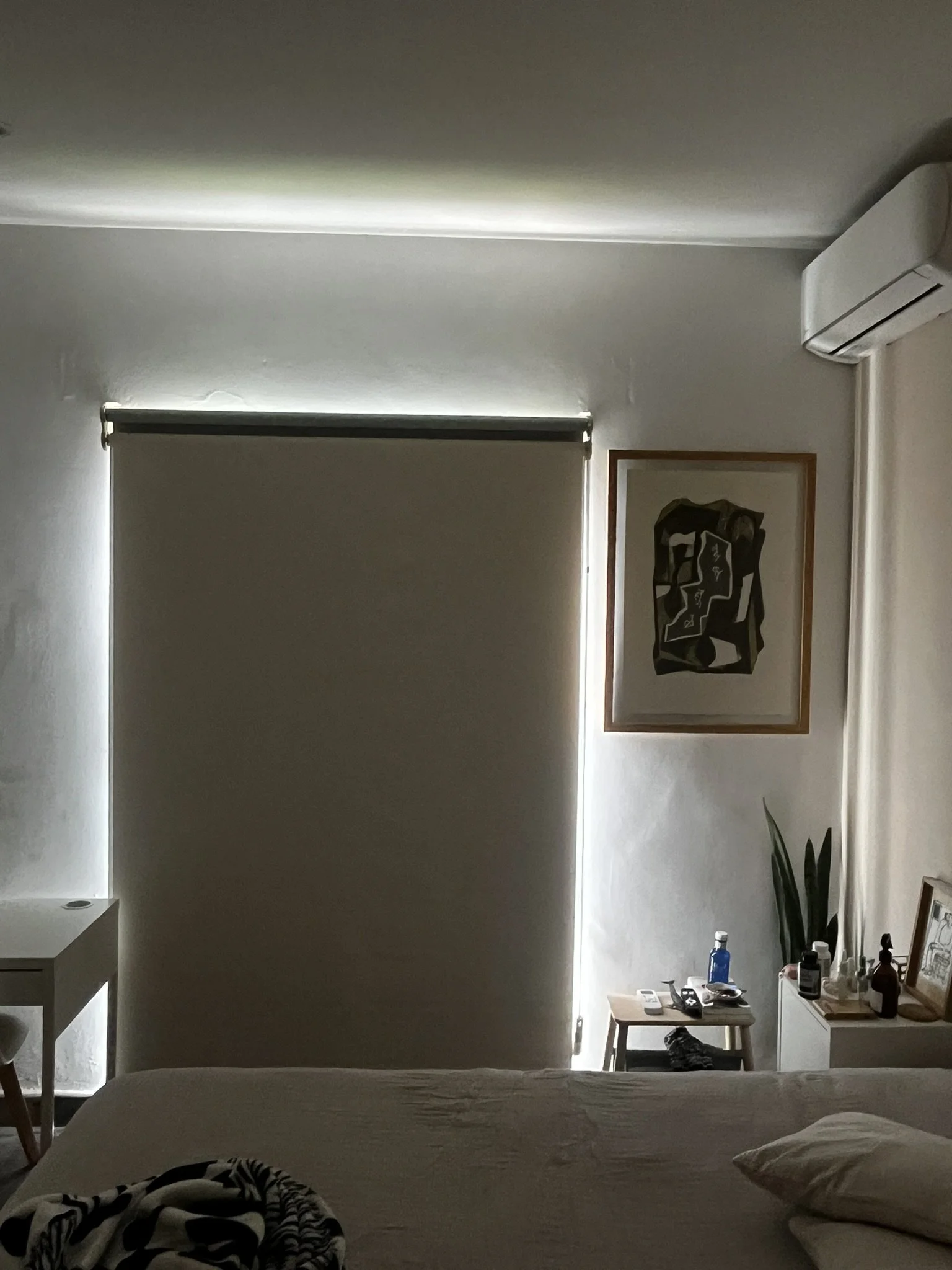Una habitación con una pared frontal con cortinas blancas, una mesa pequeña con objetos y bebidas, y una cama en primer plano con una funda gris y almohadas blancas.