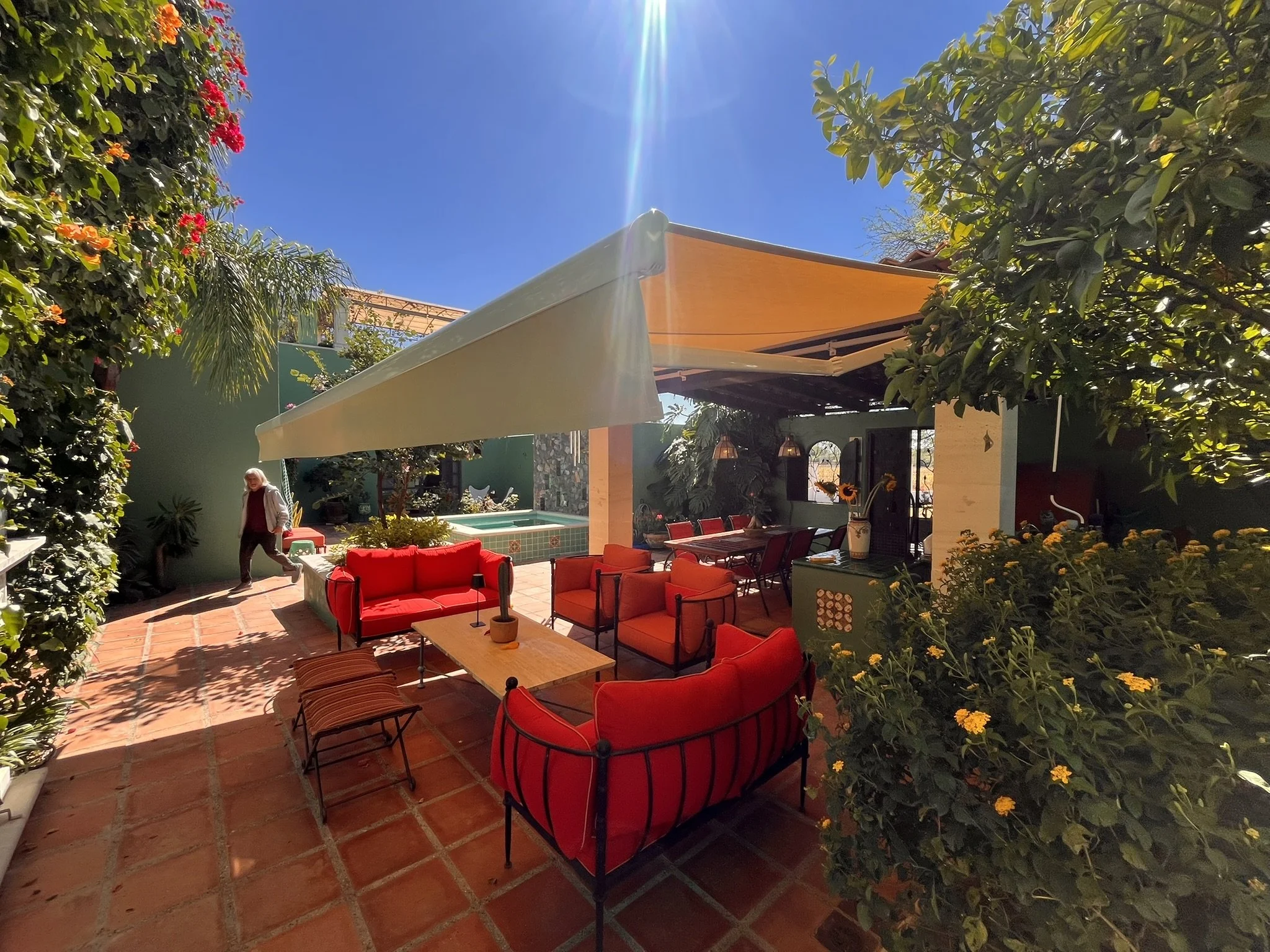 Un patio con muebles rojos y verdes, rodeado de muchas plantas y flores, con una piscina pequeña al fondo y un cielo despejado con mucho sol y un toldo retractil que protege del sol.