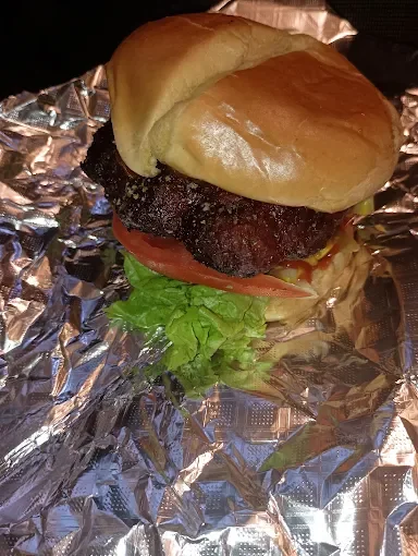 Close-up of a hamburger with a bun, lettuce, tomato, a beef patty, and a top bun, wrapped in aluminum foil.