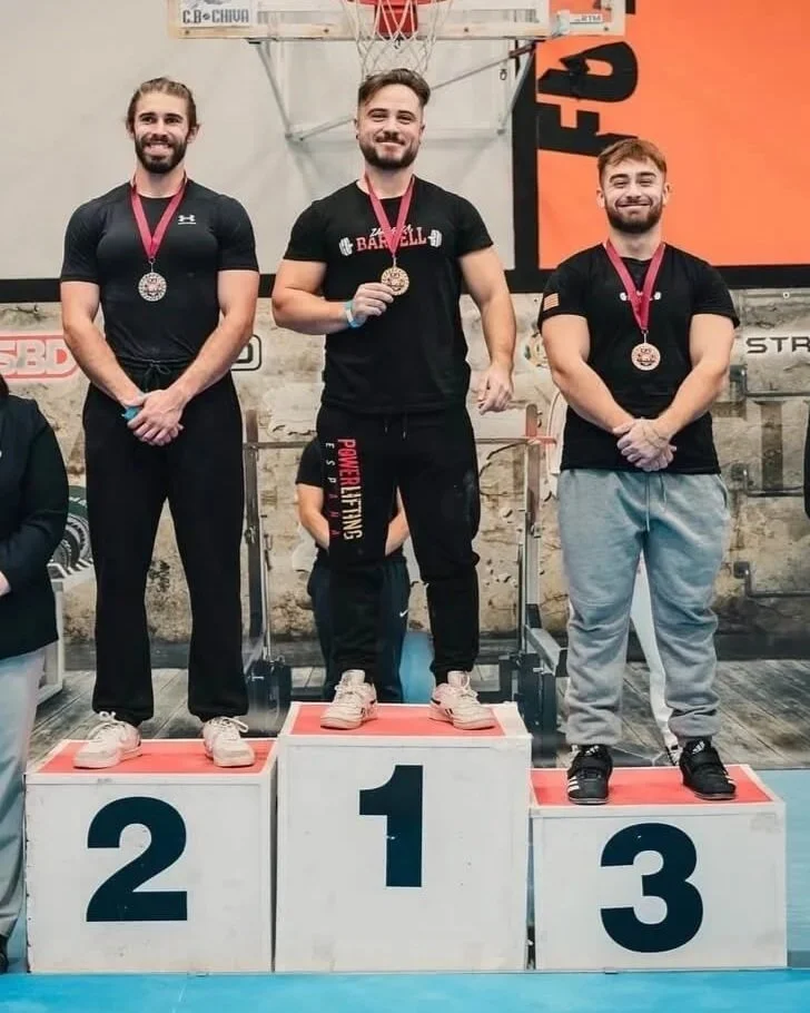 Tres hombres en un podio de competencia, con medallas alrededor del cuello, en un gimnasio de levantamiento de pesas o crossfit, celebrando su podium en el primer lugar, segundo y tercer lugar.