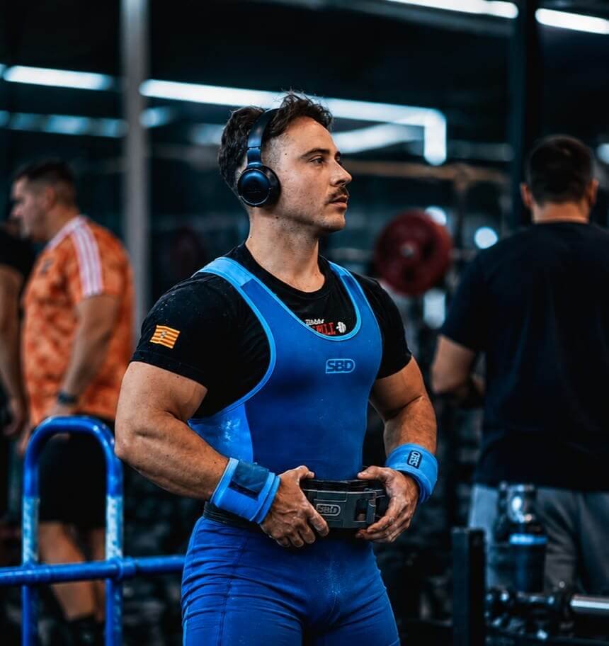 Un hombre en un gimnasio con sandalias, vestido con ropa de entrenamiento en azul y negro, usando audífonos y sosteniendo una pelota de baloncesto. En el fondo se ven otras personas entrenando y equipo de pesas.