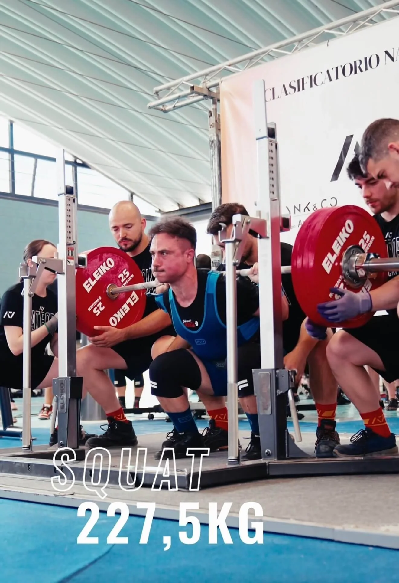Grupo de deportistas en una competencia de halterofilia en un gimnasio cubierto, preparándose para levantar pesas con una barra de 227.5 kg, con un cartel que indica 'Squat 227.5 kg'.