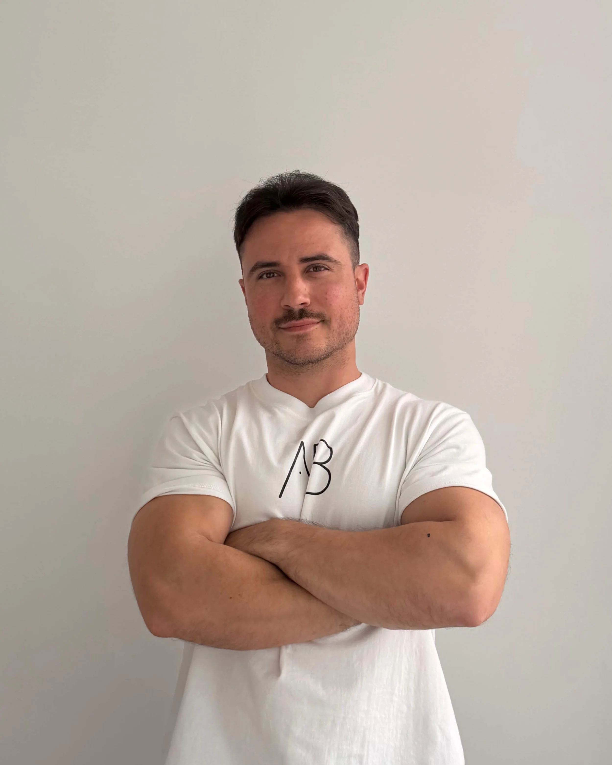 Hombre con brazos cruzados, con peinado corto y barba de dos días, viste una camiseta blanca con letras. Fondo liso y color claro.